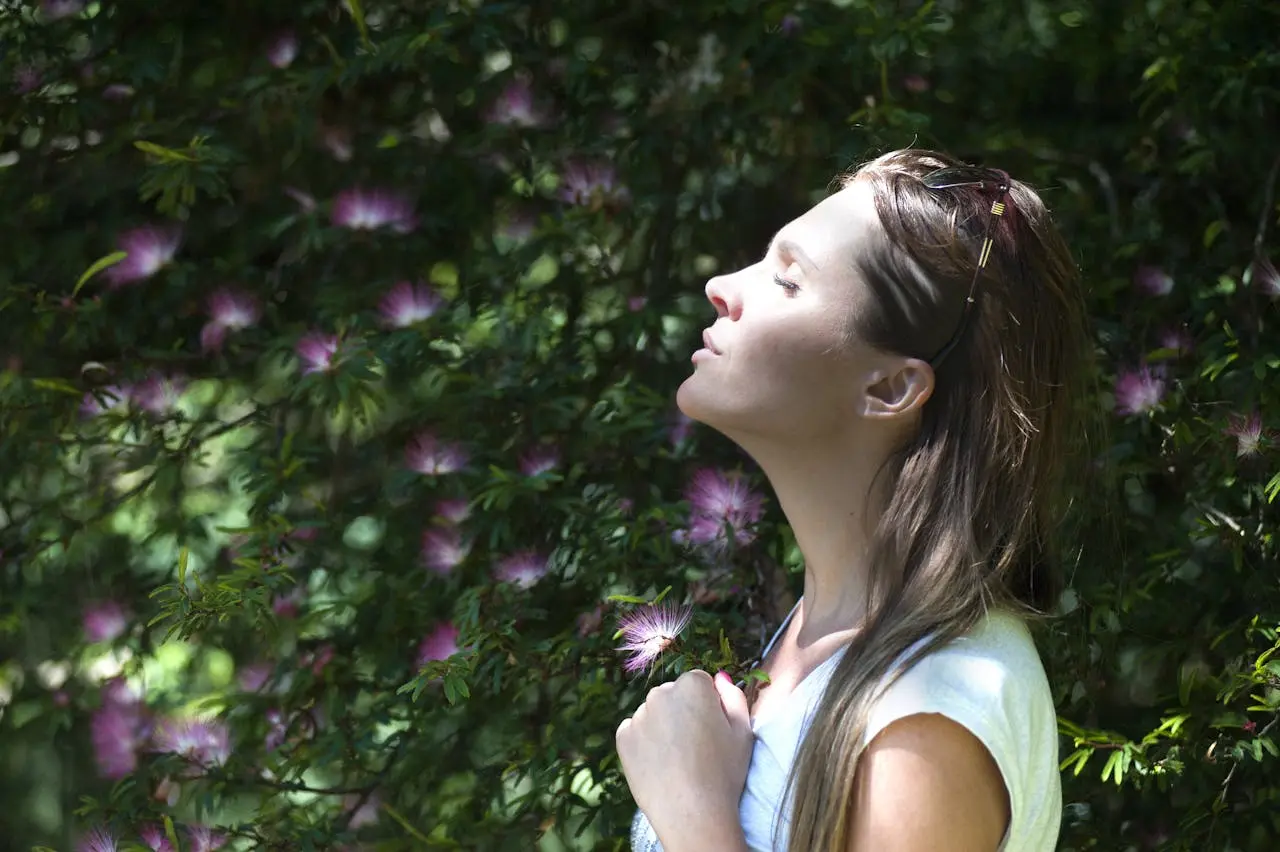 Woman standing near purple-petaled flower, closing eyes against sunlight.