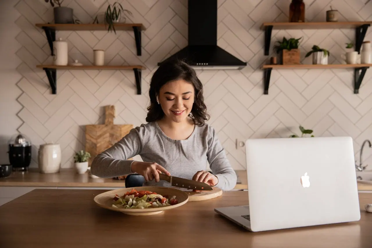 woman-slicing-food-on-a-kitchen-table-while-using-her-computer