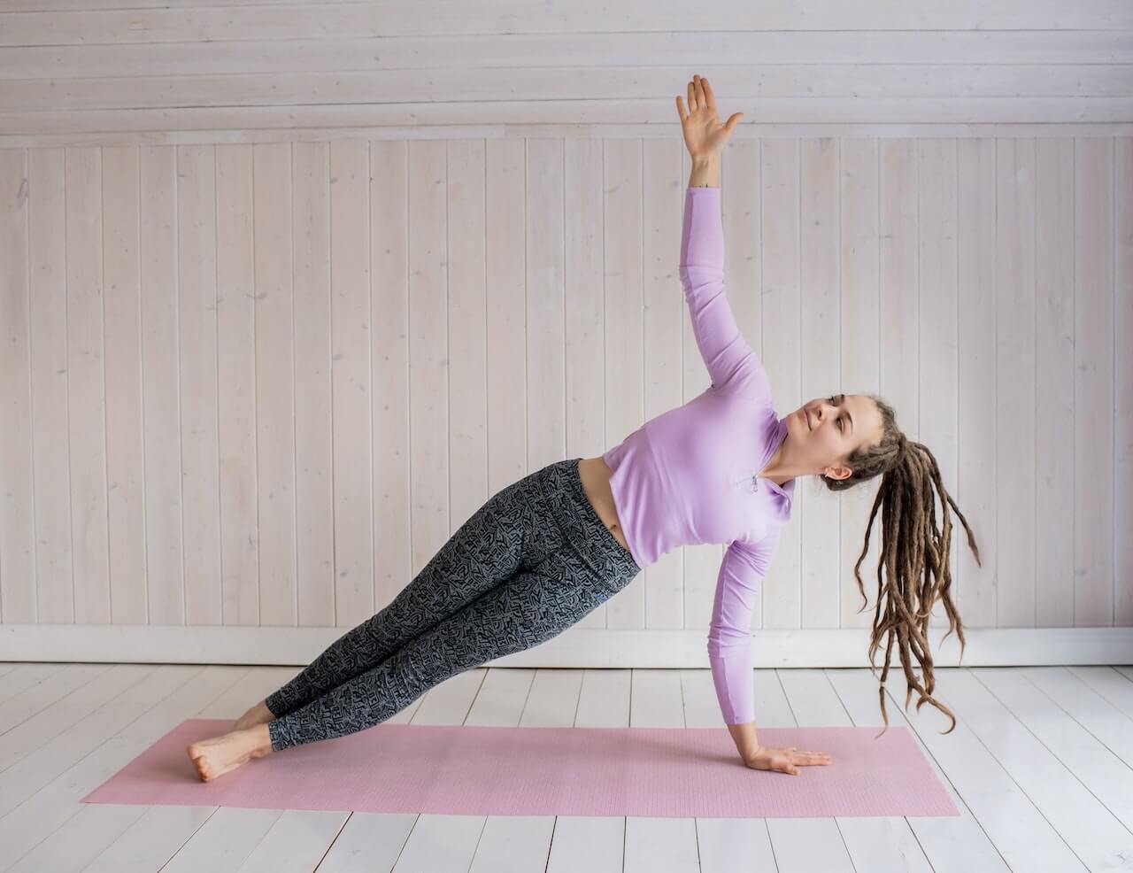 Woman in purple shirt performing side plank yoga pose on pink mat indoors