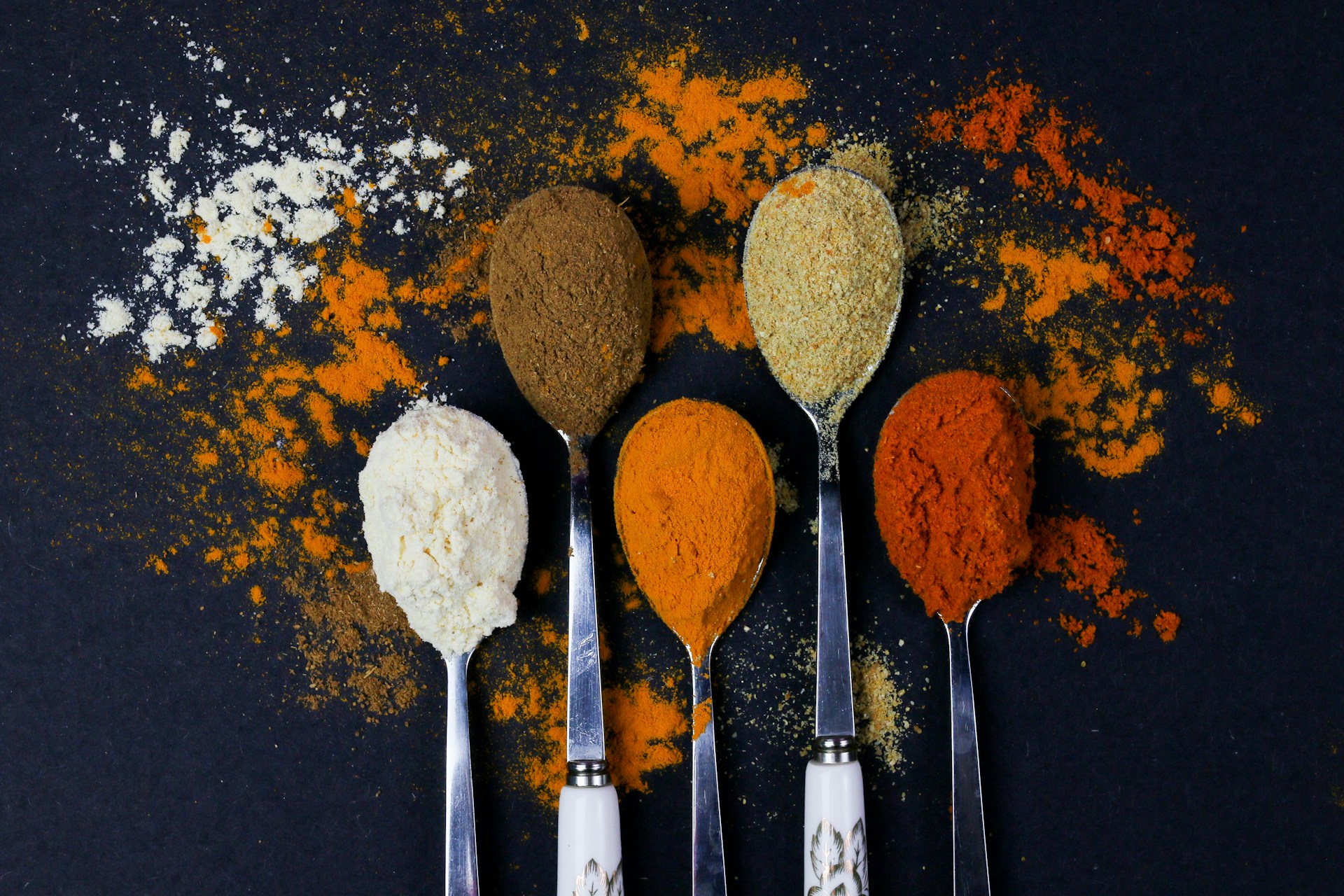 Colorful spices in spoons with scattered powder on dark background