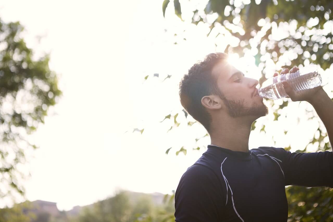 Person drinking water from bottle outdoors with sunlight filtering through trees