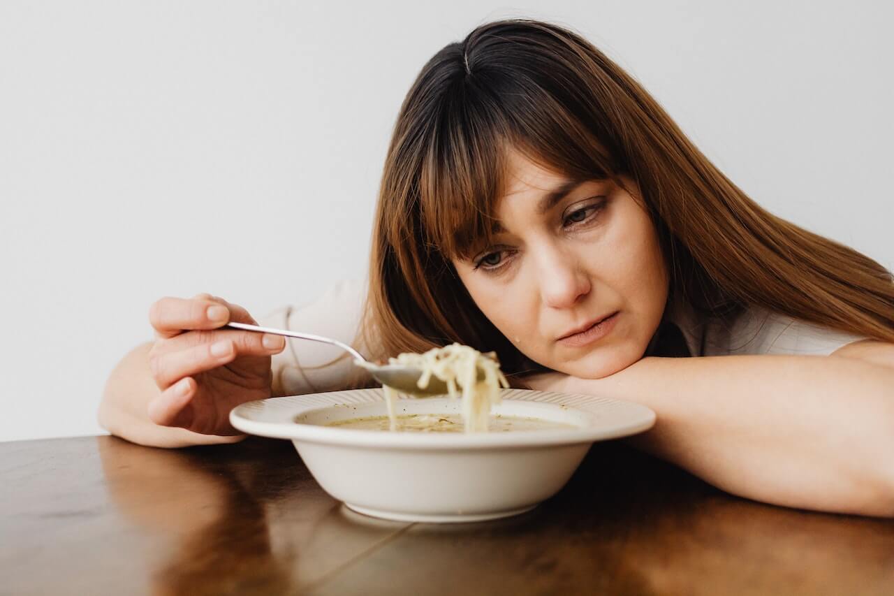 Tired woman eating soup while leaning on table, looking unenthusiastic
