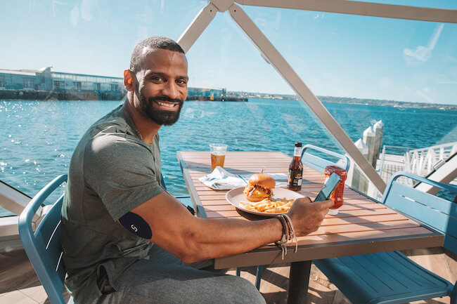 Smiling man enjoying burger and beer at waterfront table with ocean view