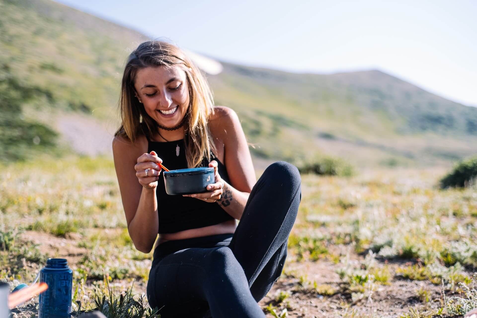 Woman smiling and eating from a bowl outdoors in grassy mountainous landscape
