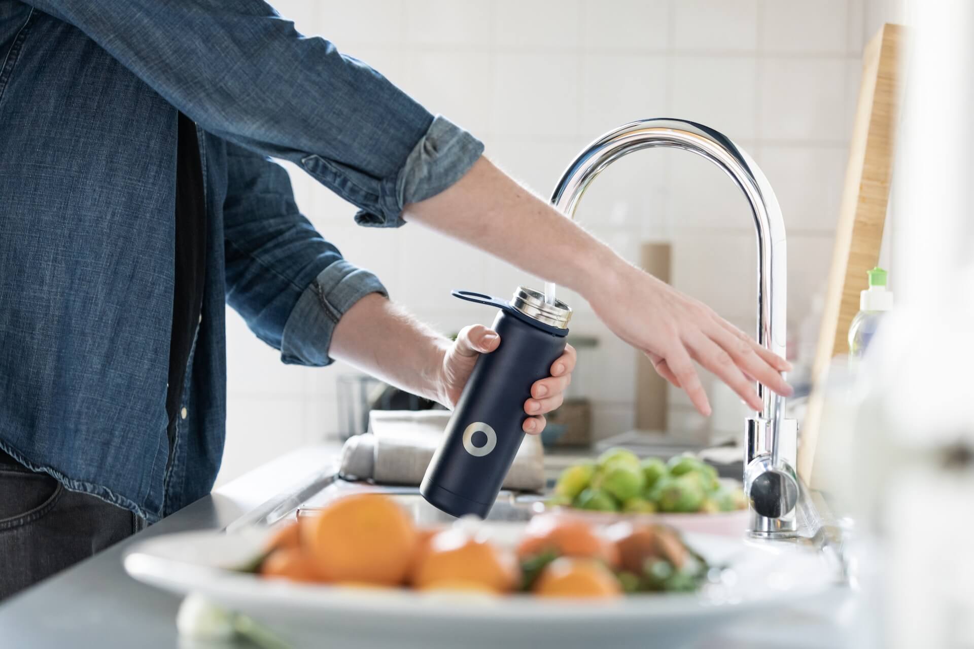 Person filling reusable water bottle at kitchen sink with fresh produce