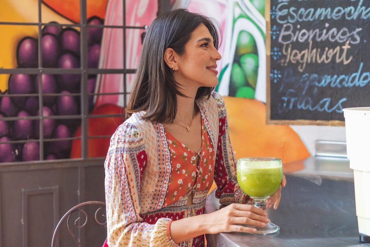 Woman in colorful dress enjoying green smoothie at cafe with fruit backdrop