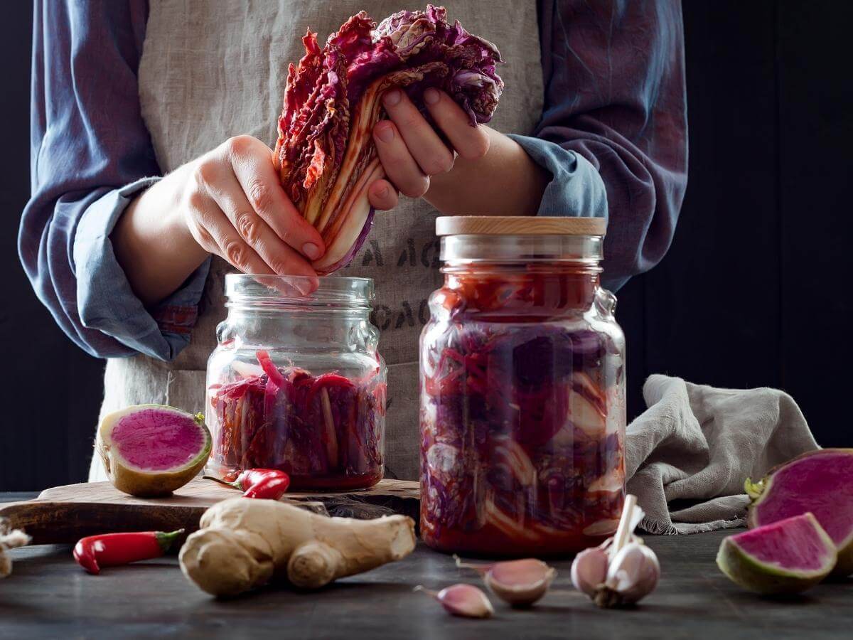 Hands packing fermented red cabbage into glass jars with ginger and chili