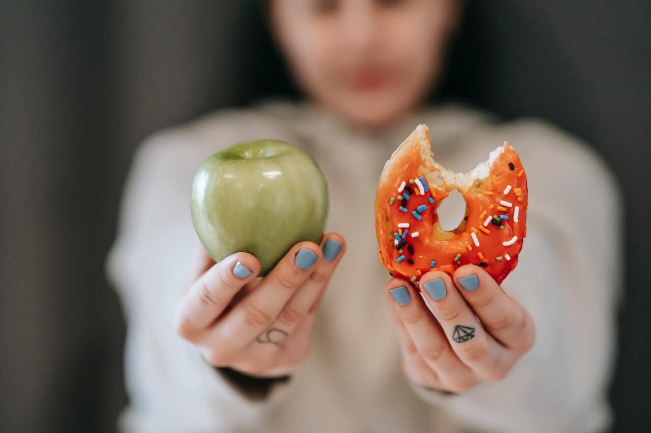 Hands holding green apple and colorful sprinkled donut with blue nails