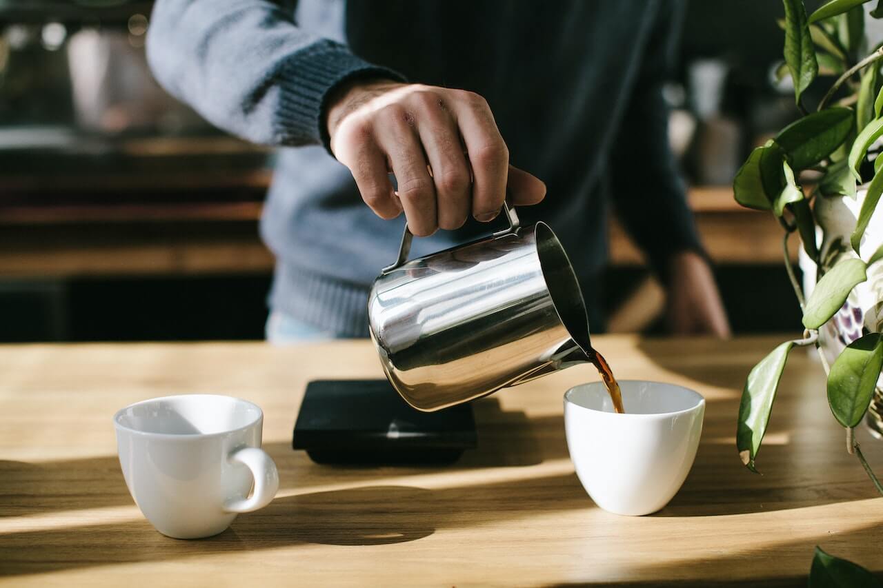 Hand pouring coffee from metal kettle into white ceramic cup