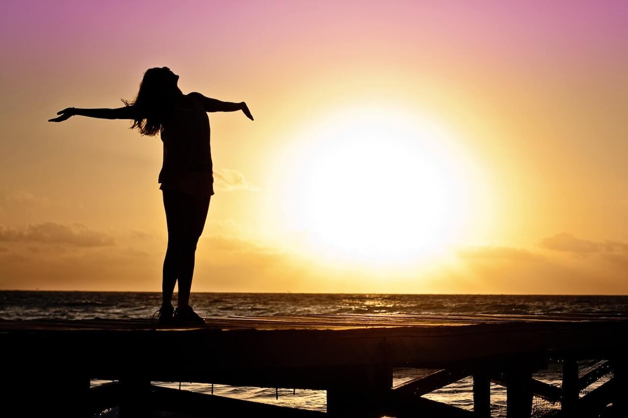 Silhouette of person with arms spread standing on pier at golden sunset