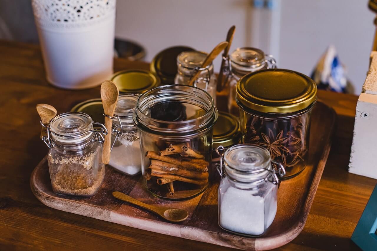 Glass jars with spices, sugar, and wooden spoons arranged on a wooden tray.