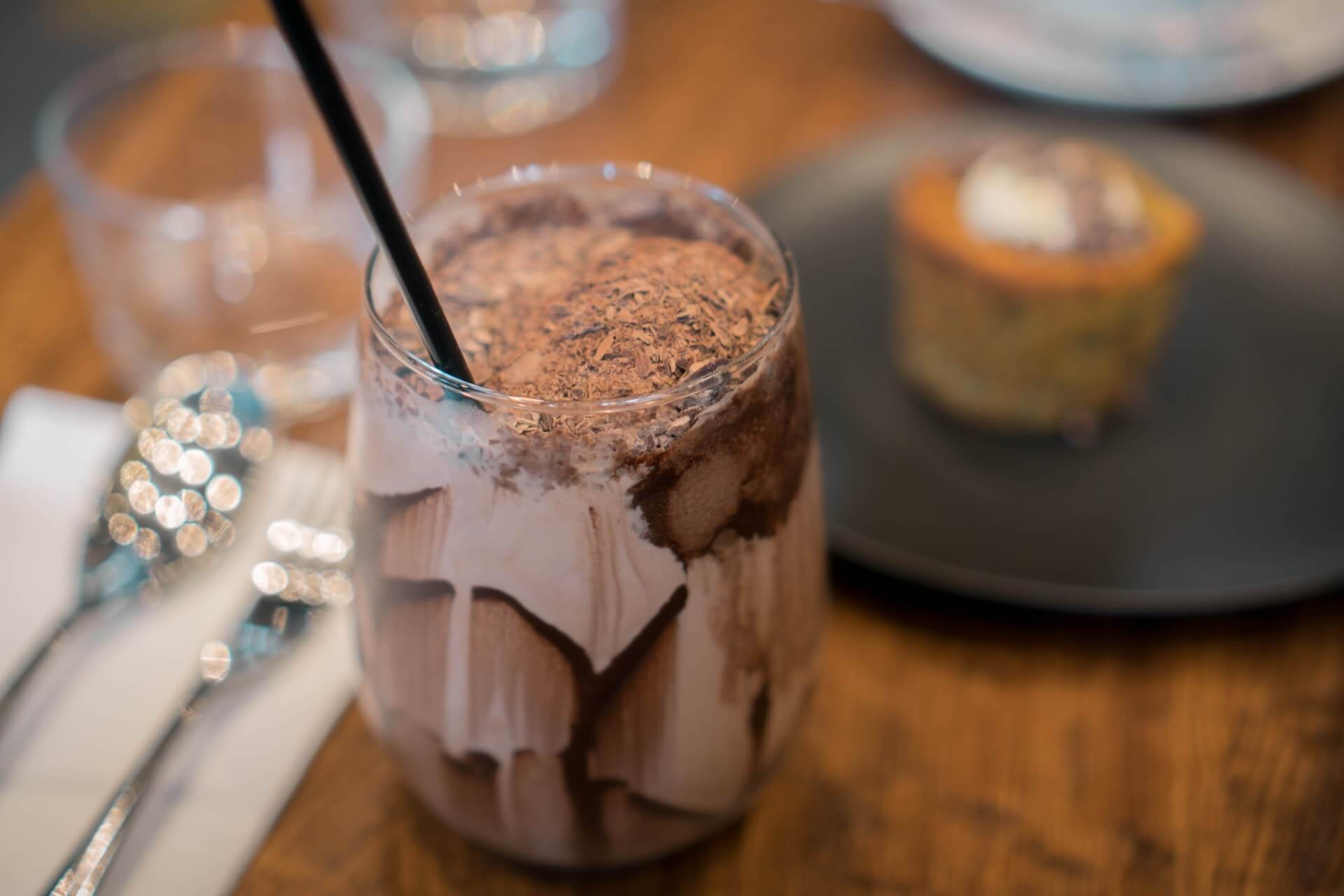 Glass of iced chocolate drink with black straw on wooden table with dinnerware