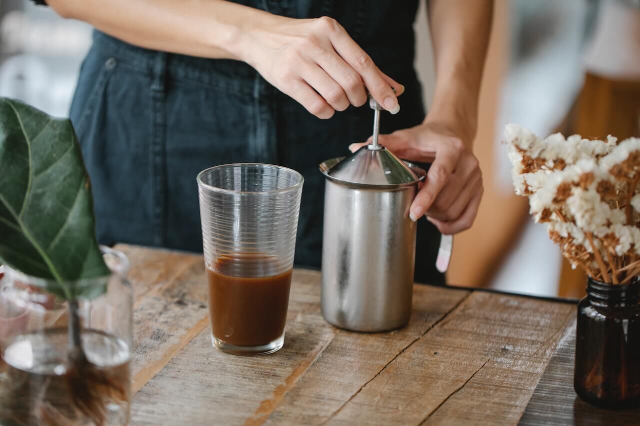 Person stirring cold coffee with metal milk frother pitcher on wooden table