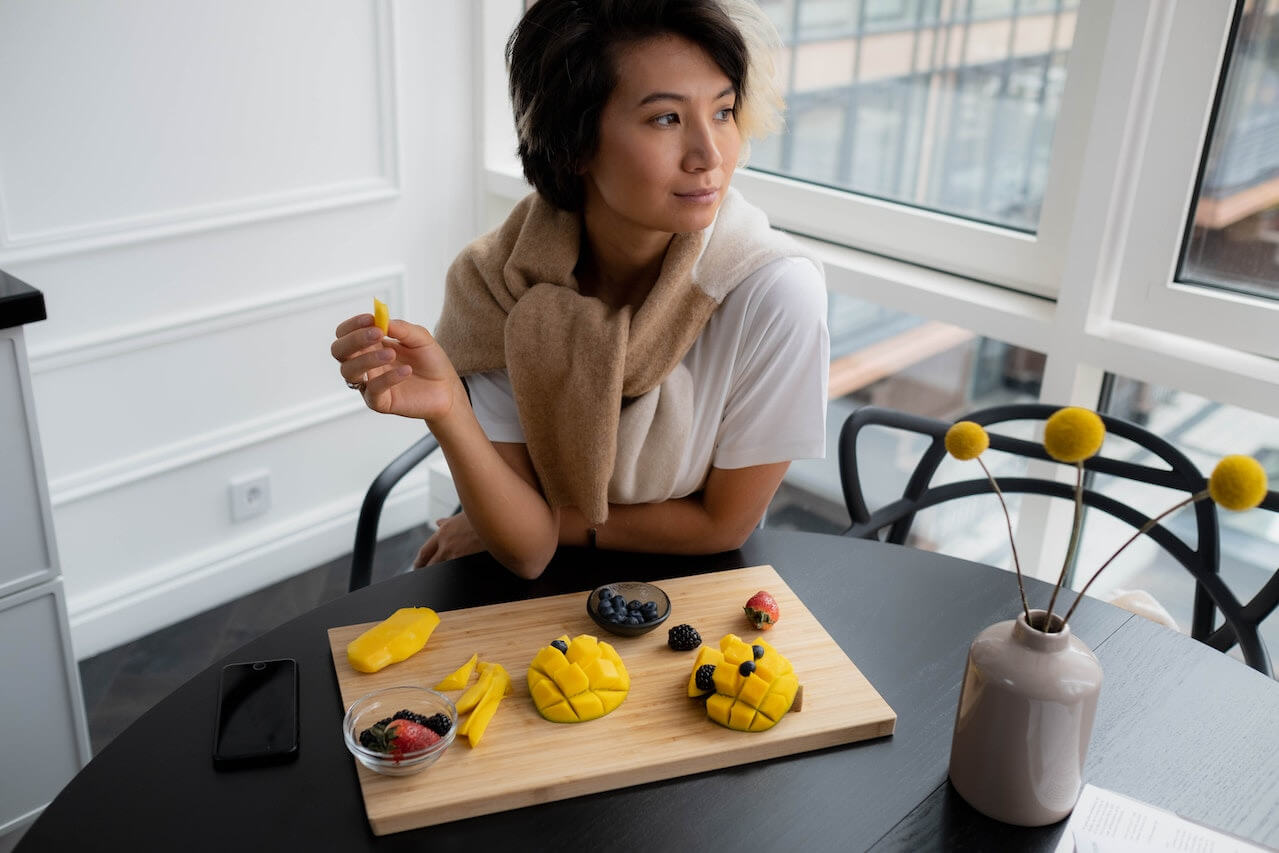 Woman holding mango slice at table with fresh fruit and yellow flowers