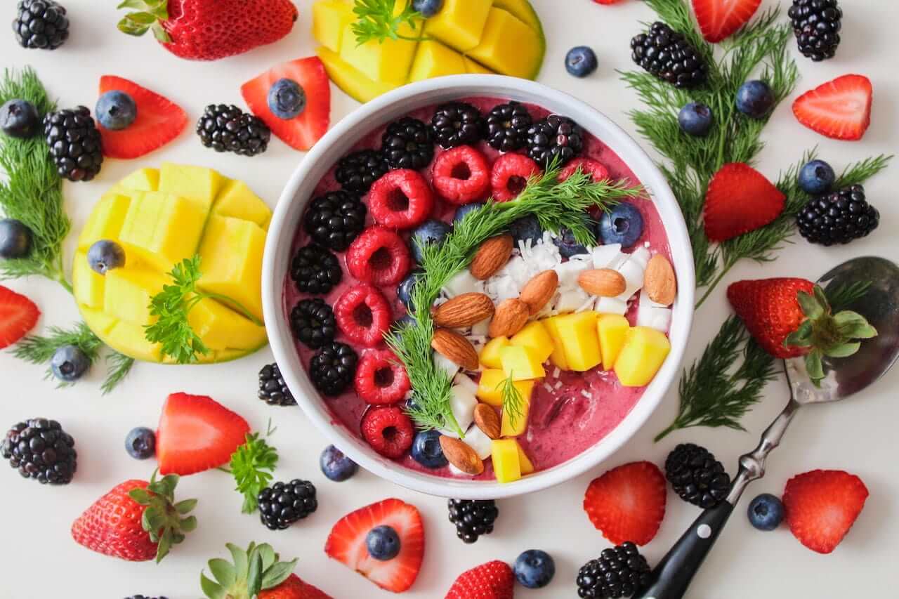 Colorful smoothie bowl topped with berries, almonds, coconut, mango, surrounded by fresh fruit.
