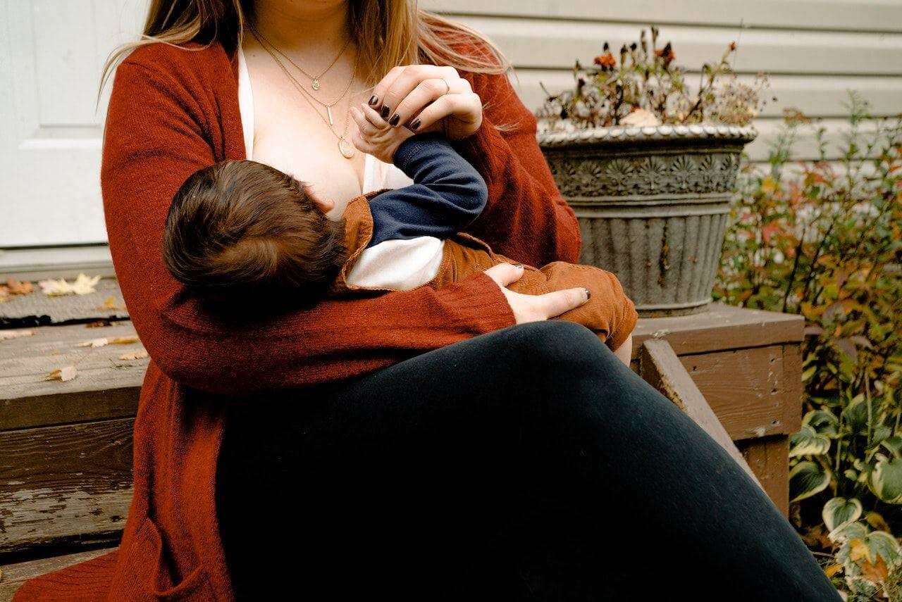 Mother in rust-colored sweater breastfeeding baby on wooden bench outdoors