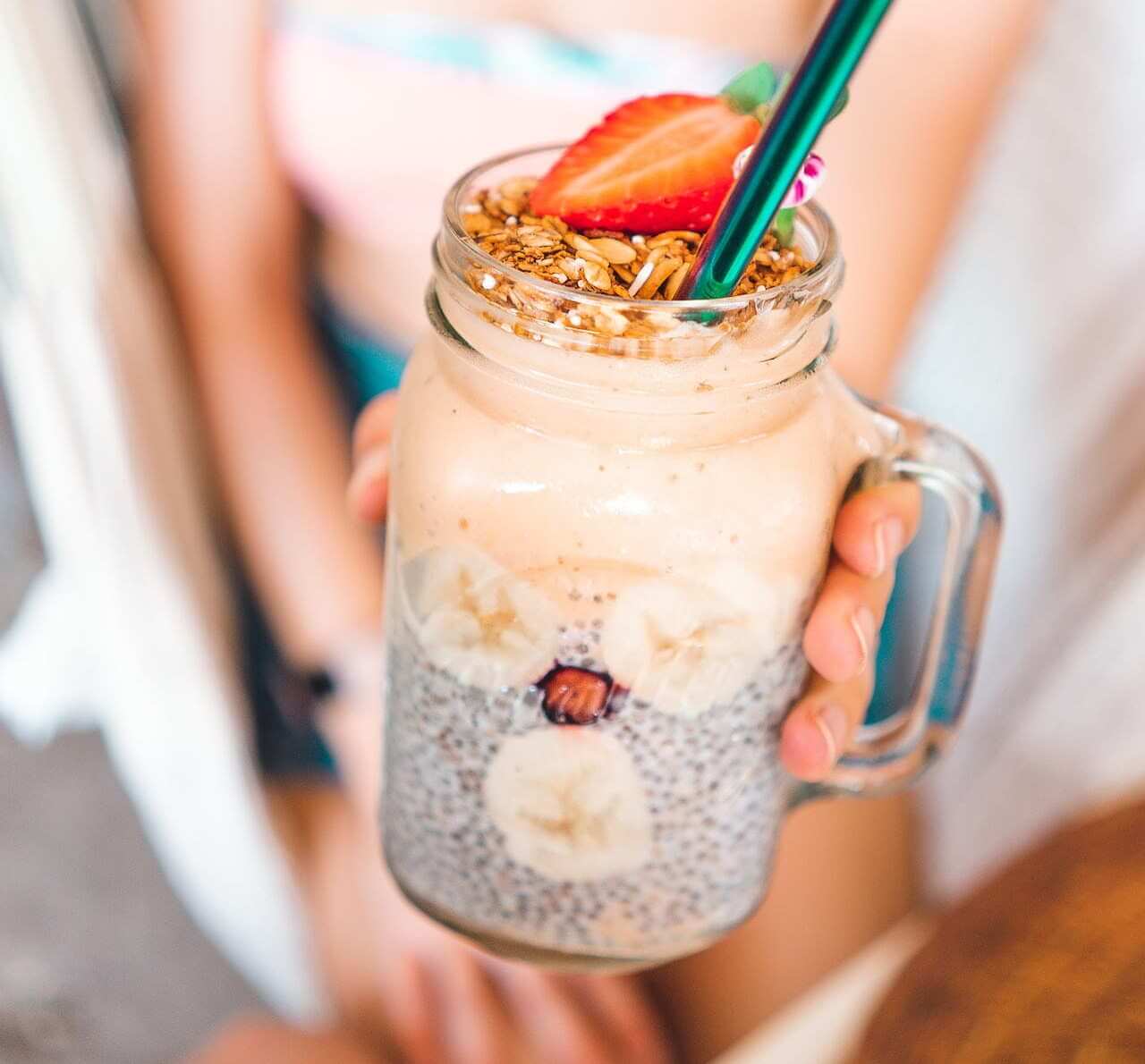 Layered chia pudding with granola, strawberry, and green straw in glass jar
