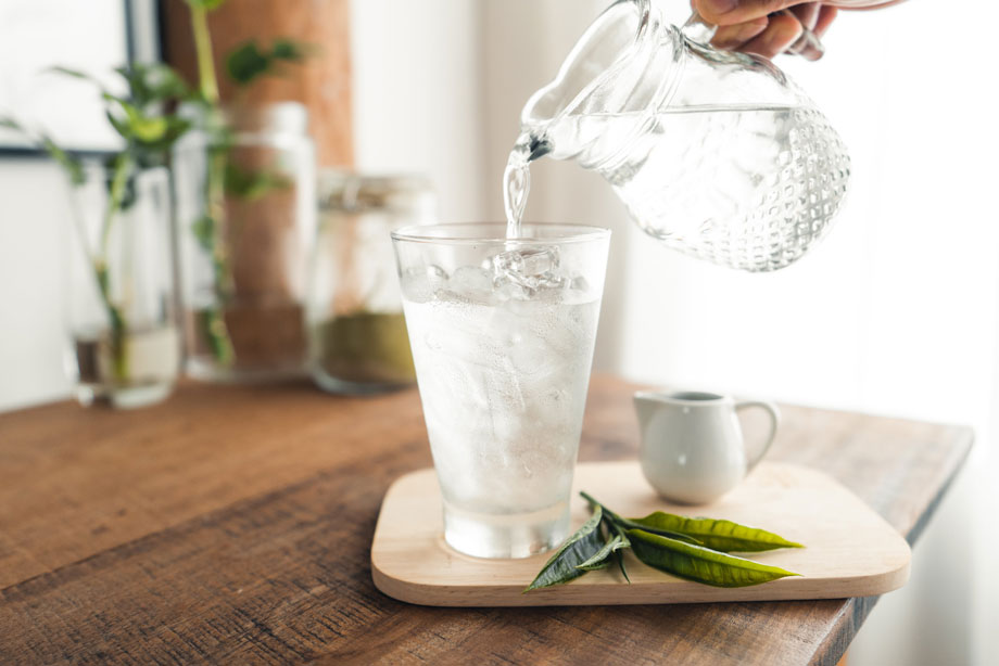 Person pouring water from glass pitcher into fresh drink with ice