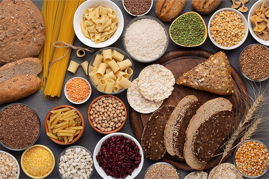 Assorted grains, legumes, pasta, bread, and seeds arranged in bowls on dark surface.