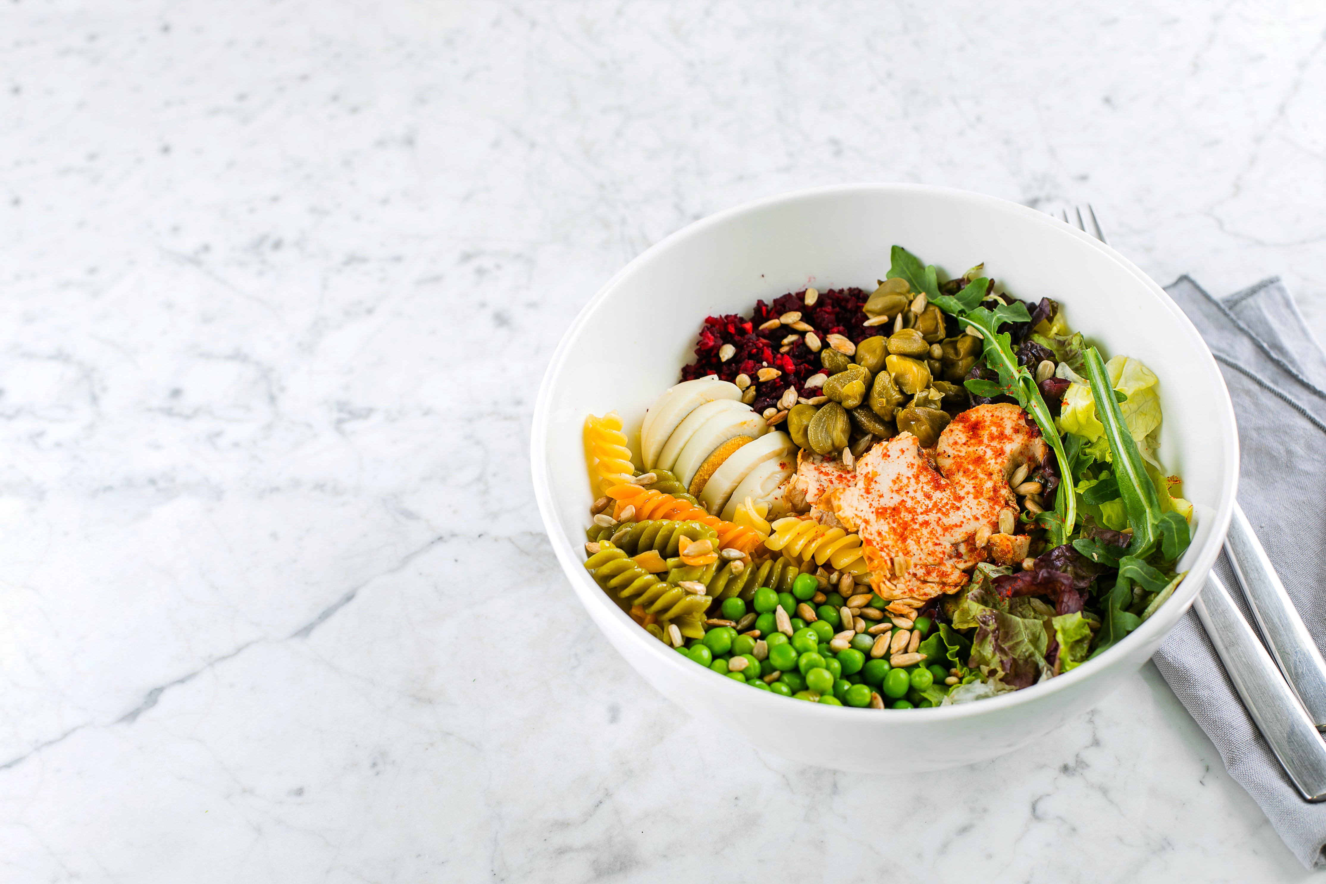 Colorful salad bowl with vegetables, pasta, greens, and protein on marble surface.