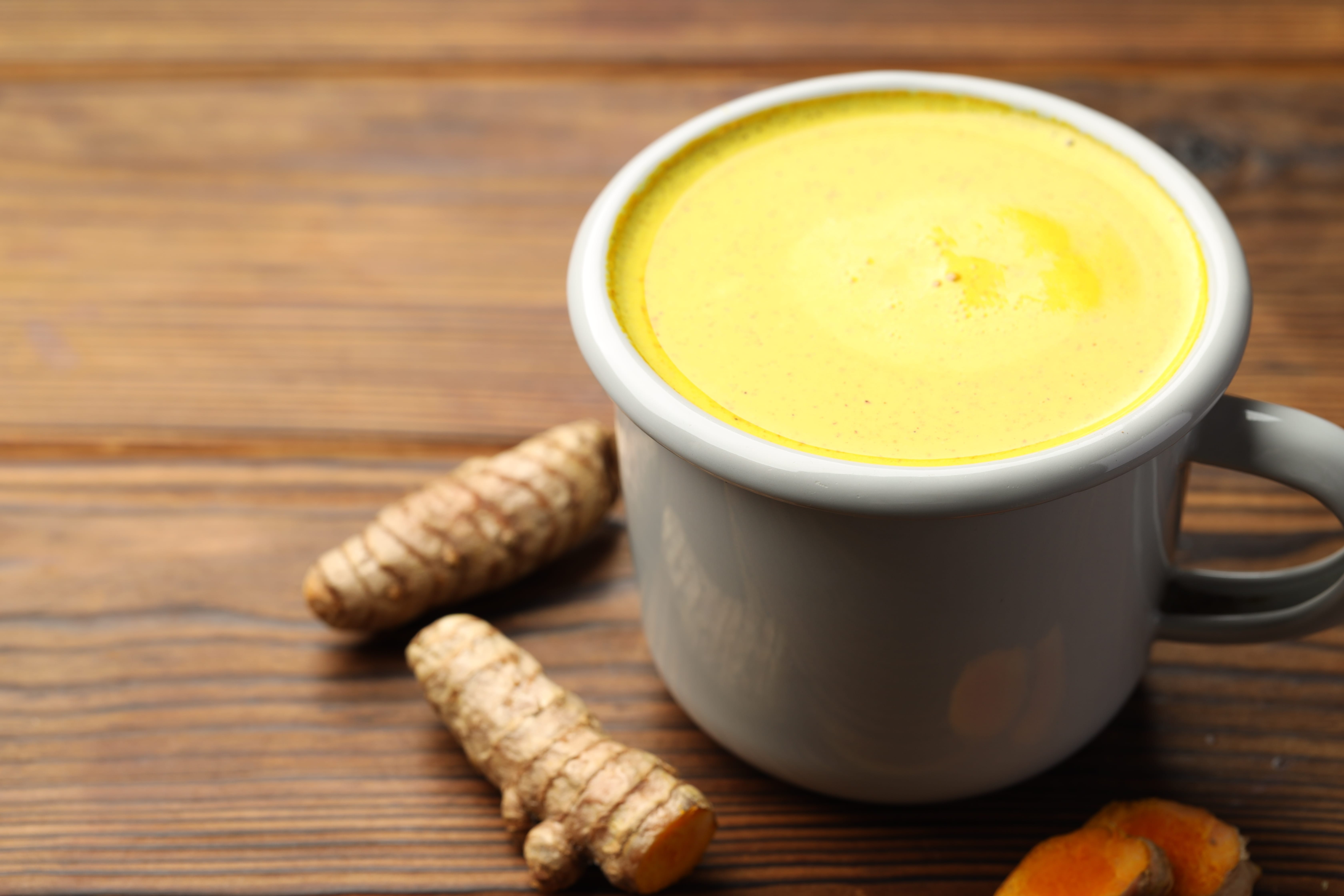 White mug of yellow turmeric drink with ginger root on wooden table