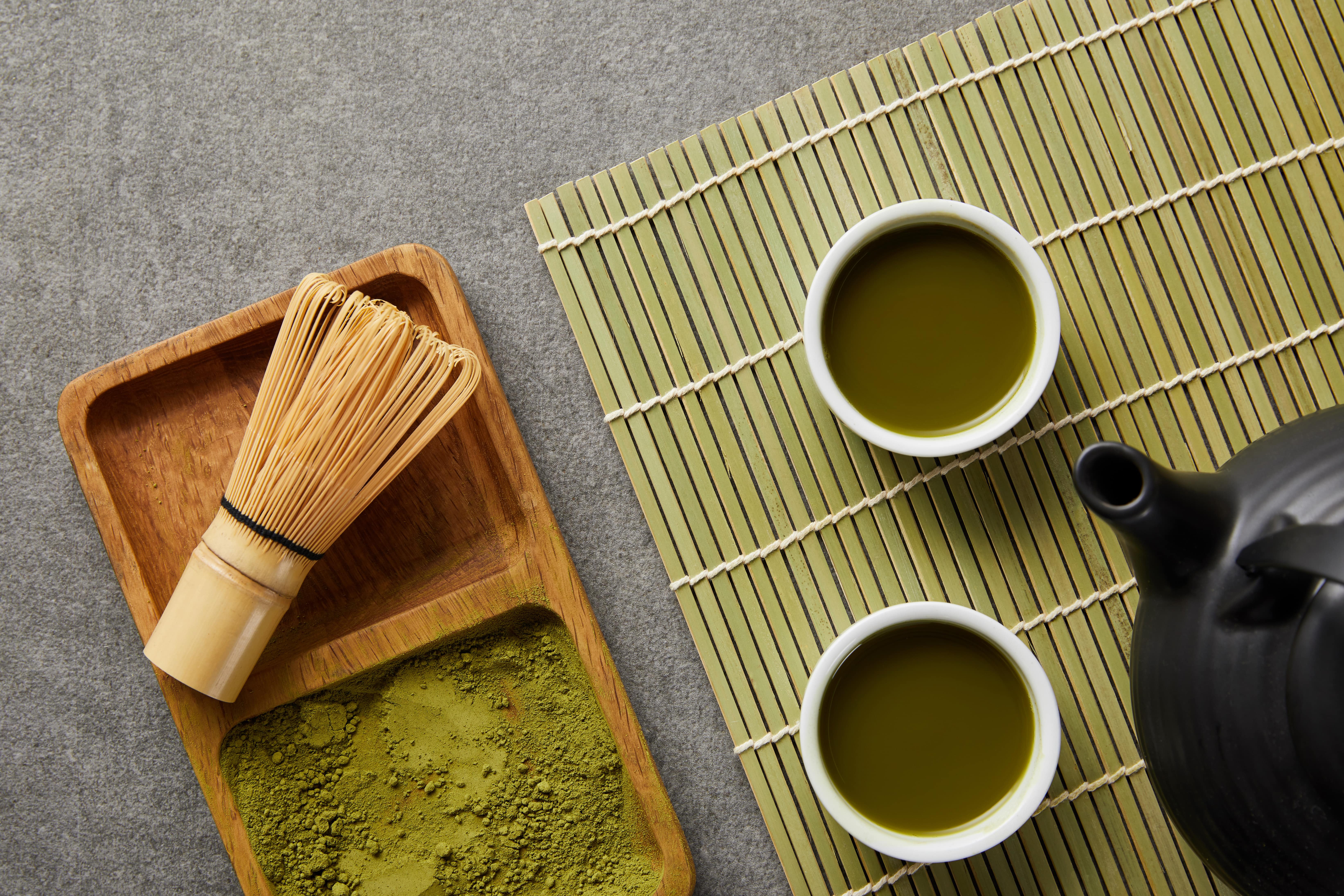Matcha tea setup with powder, whisk, bowls, and teapot on bamboo mat