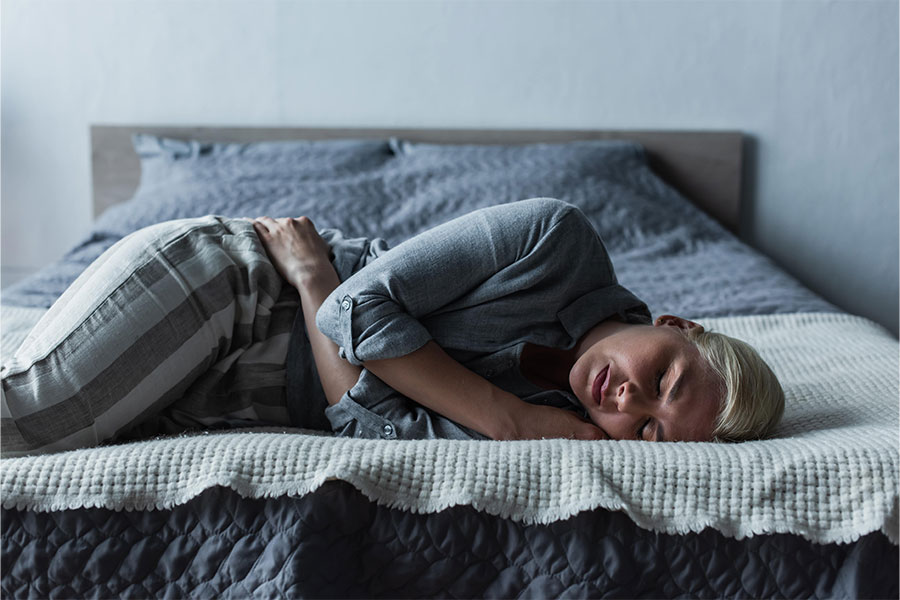 Person lying on bed with abdominal pain, holding stomach, wearing blue shirt