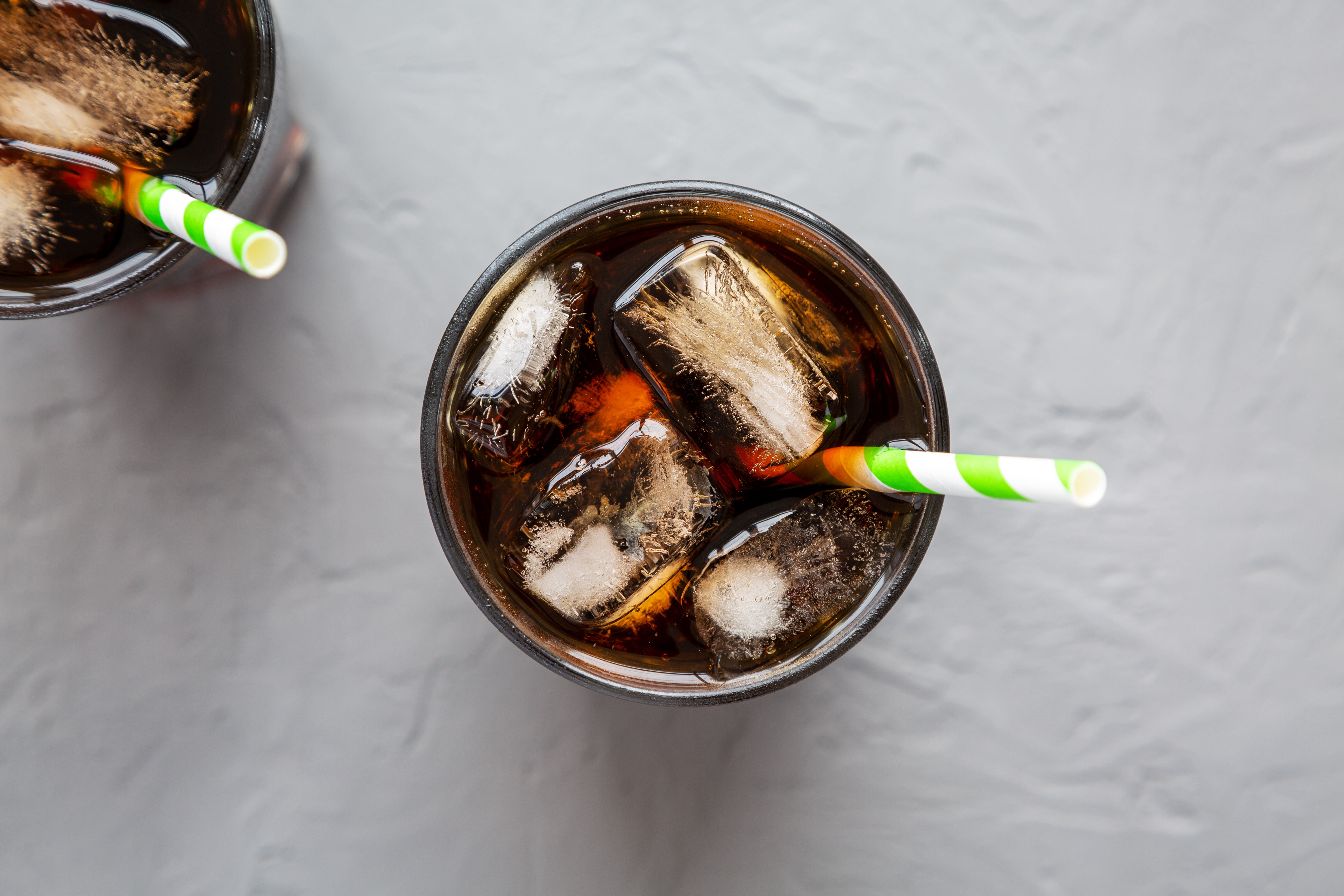 Cola drink being poured into glass with ice and striped straw on marble surface
