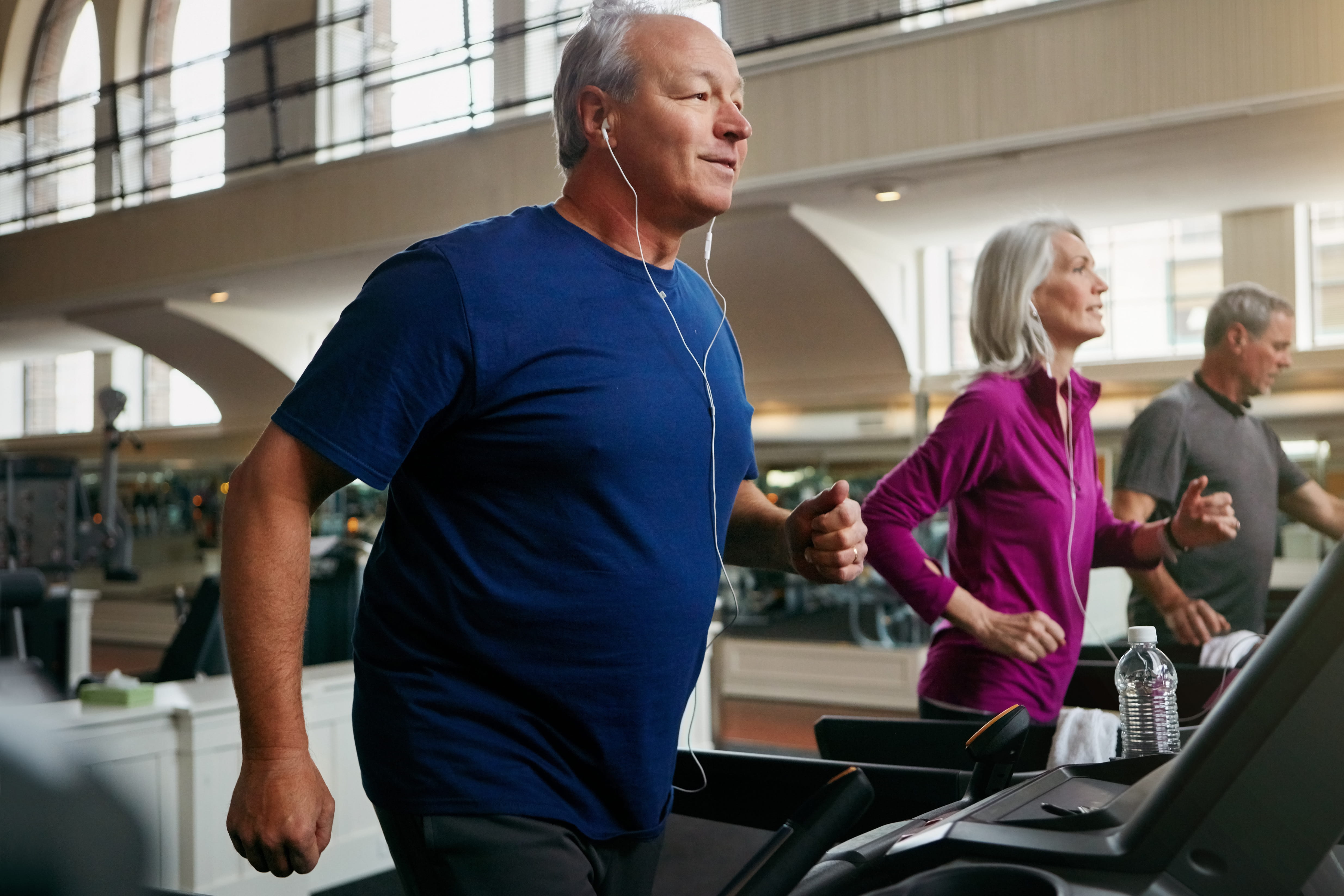 Older man in blue shirt running on treadmill with earbuds in modern gym facility