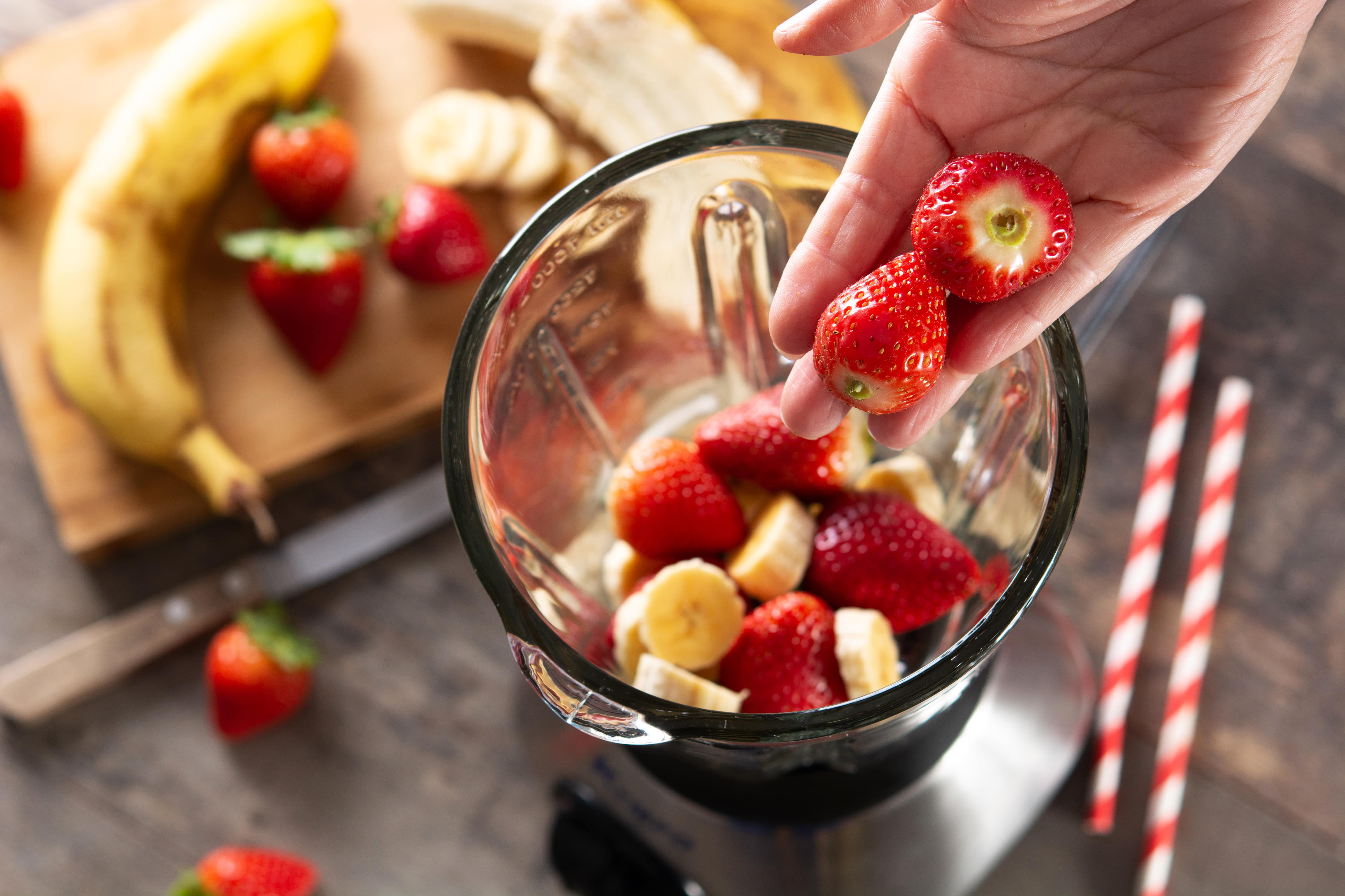 Hand adding fresh strawberries to smoothie bowl with banana and fruit ingredients