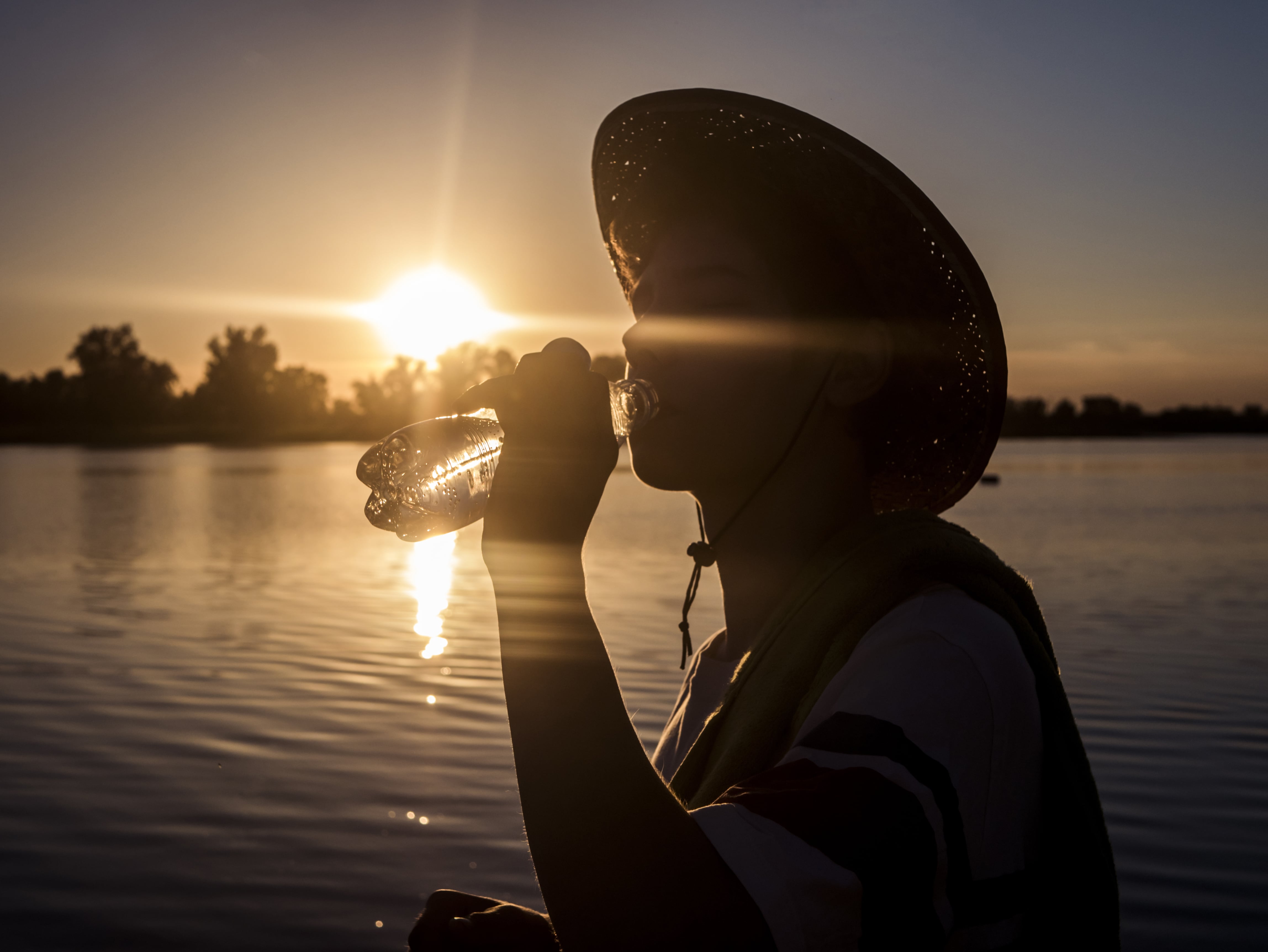 Silhouetted person in hood drinks water by riverside at sunset with trees in background.