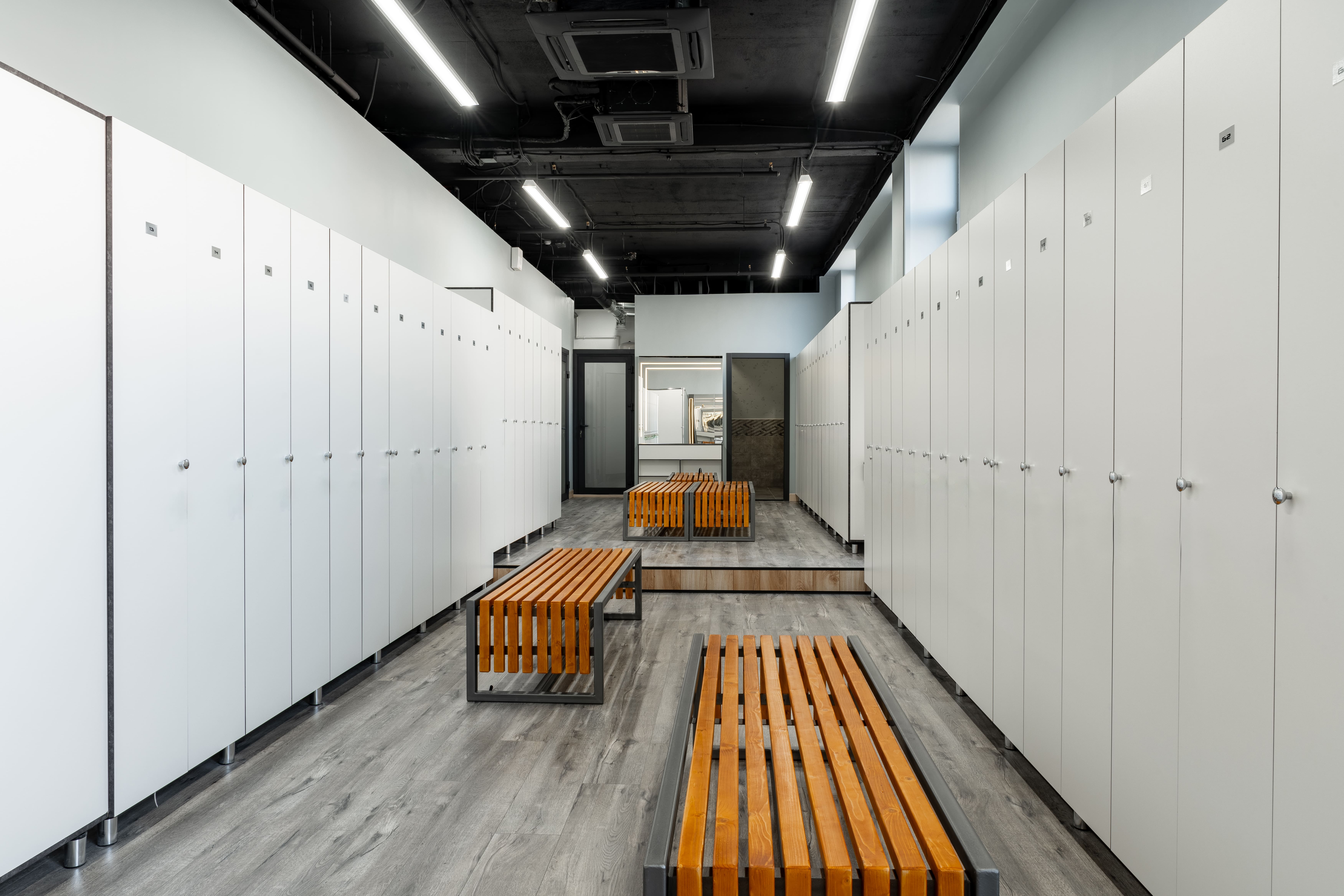 Modern locker room with white lockers, orange wooden benches, and bright fluorescent ceiling lights.