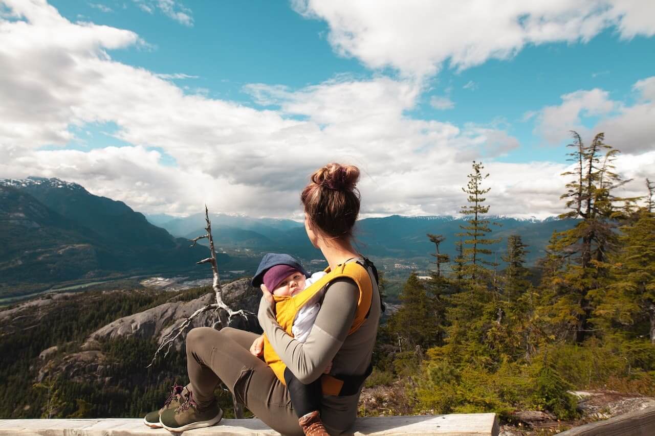 Woman hiker with toddler on back overlooks mountain valley landscape
