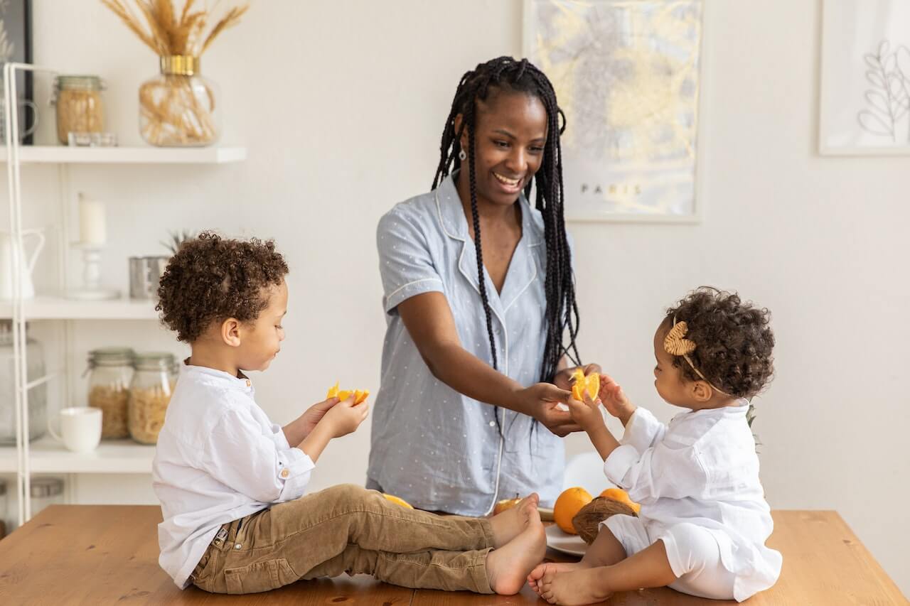 Woman sharing oranges with two young children at kitchen table in bright home.
