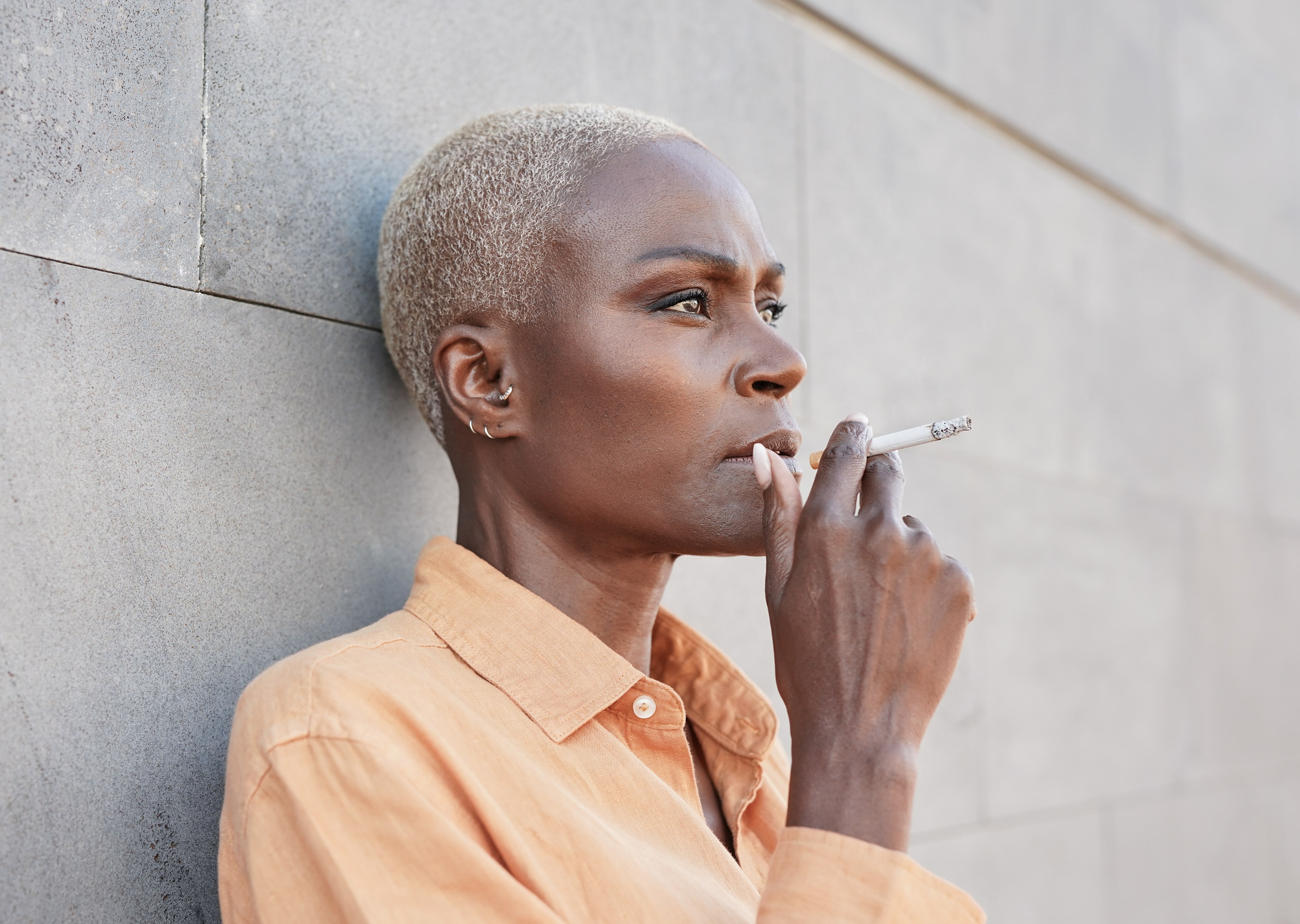 Person with short blonde hair wearing peachy shirt smoking cigarette against concrete wall
