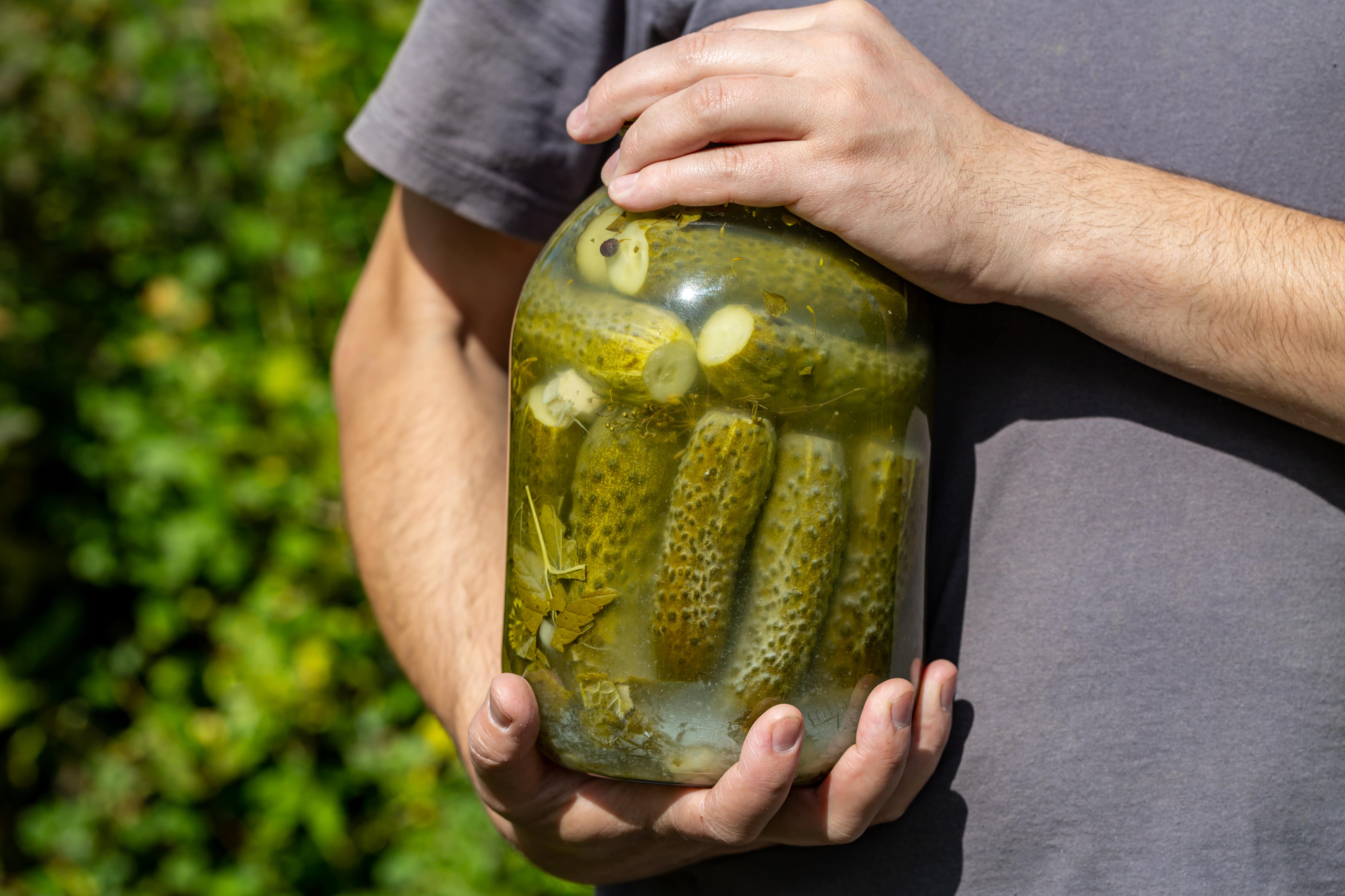Person holding a glass jar of pickles and garlic slices outdoors.