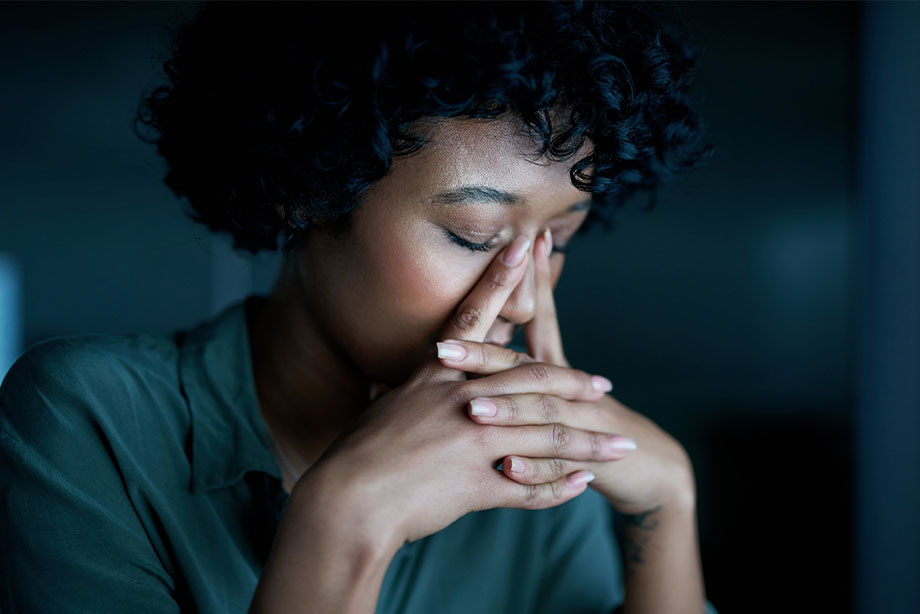 Woman with curly hair in contemplative pose, hand to face, dark clothing against blue background.