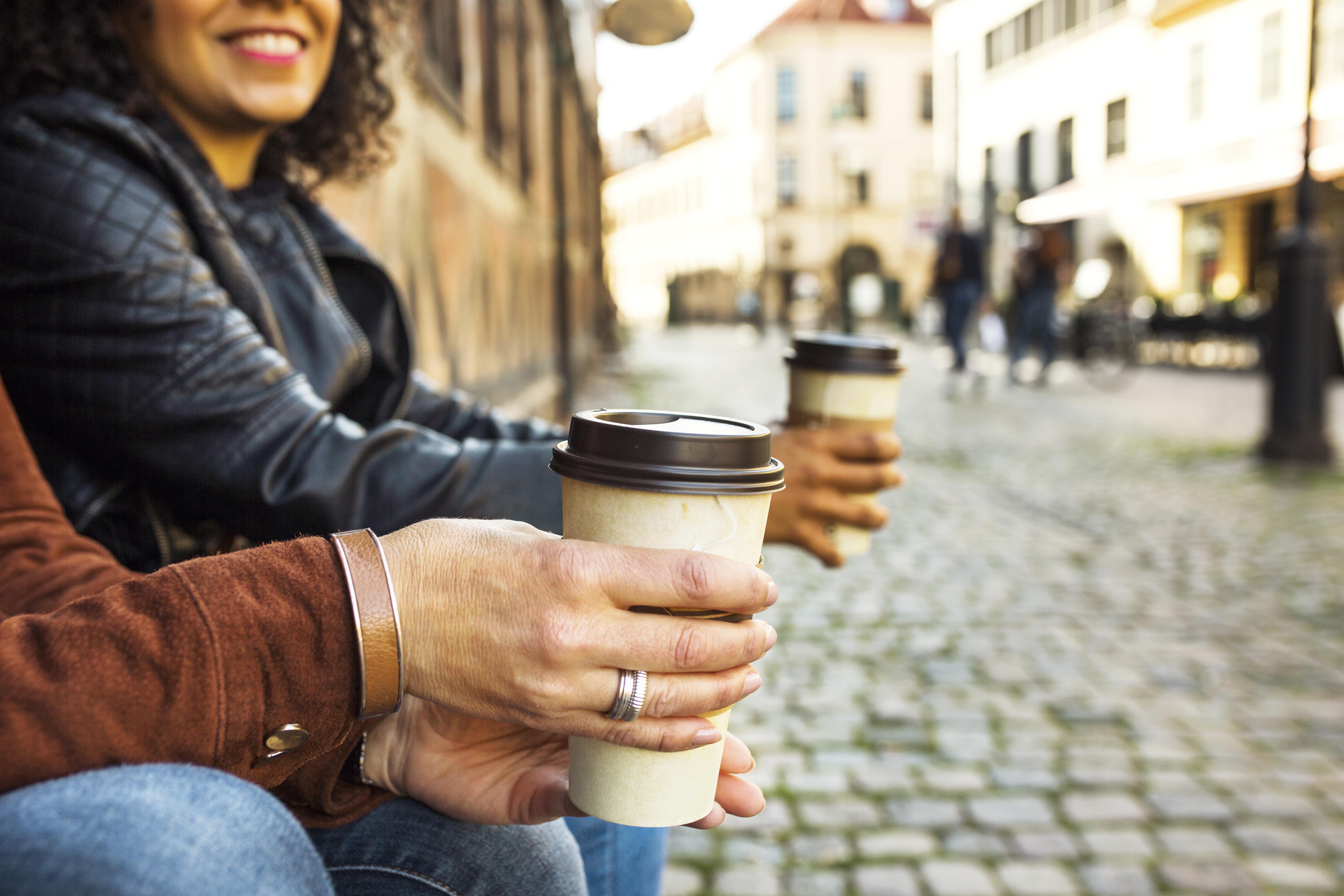 Woman holding coffee cup smiling on historic cobblestone street with buildings.