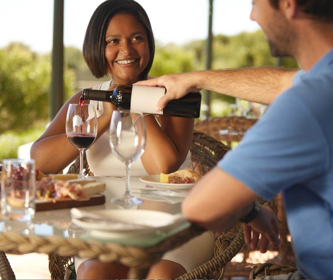 Man pours wine for smiling woman at outdoor dining table with food and nature background