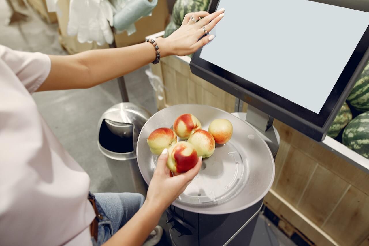 Person holding red apple at food weighing scale next to tablet screen