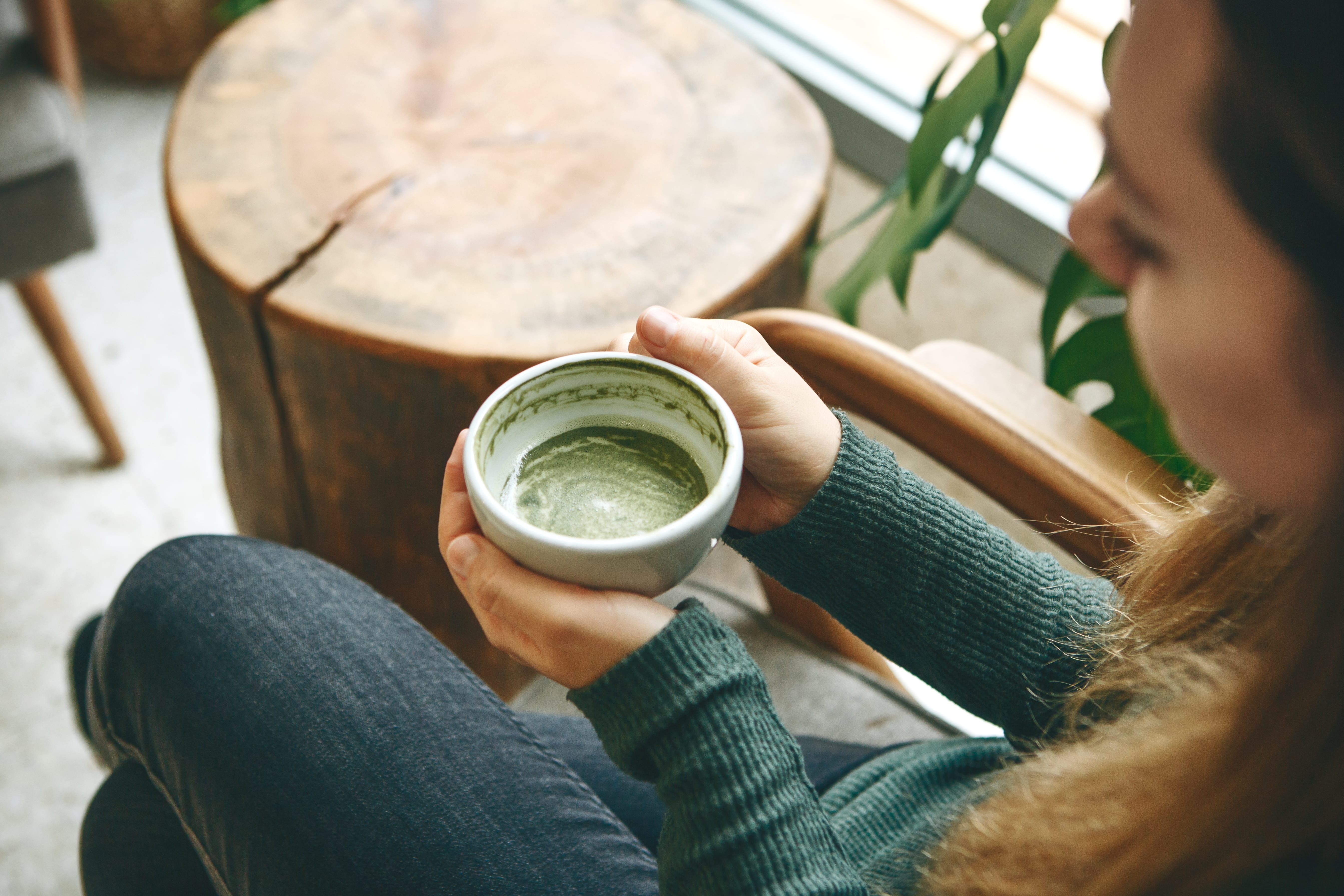 Woman holding matcha tea cup near wooden table and green plant
