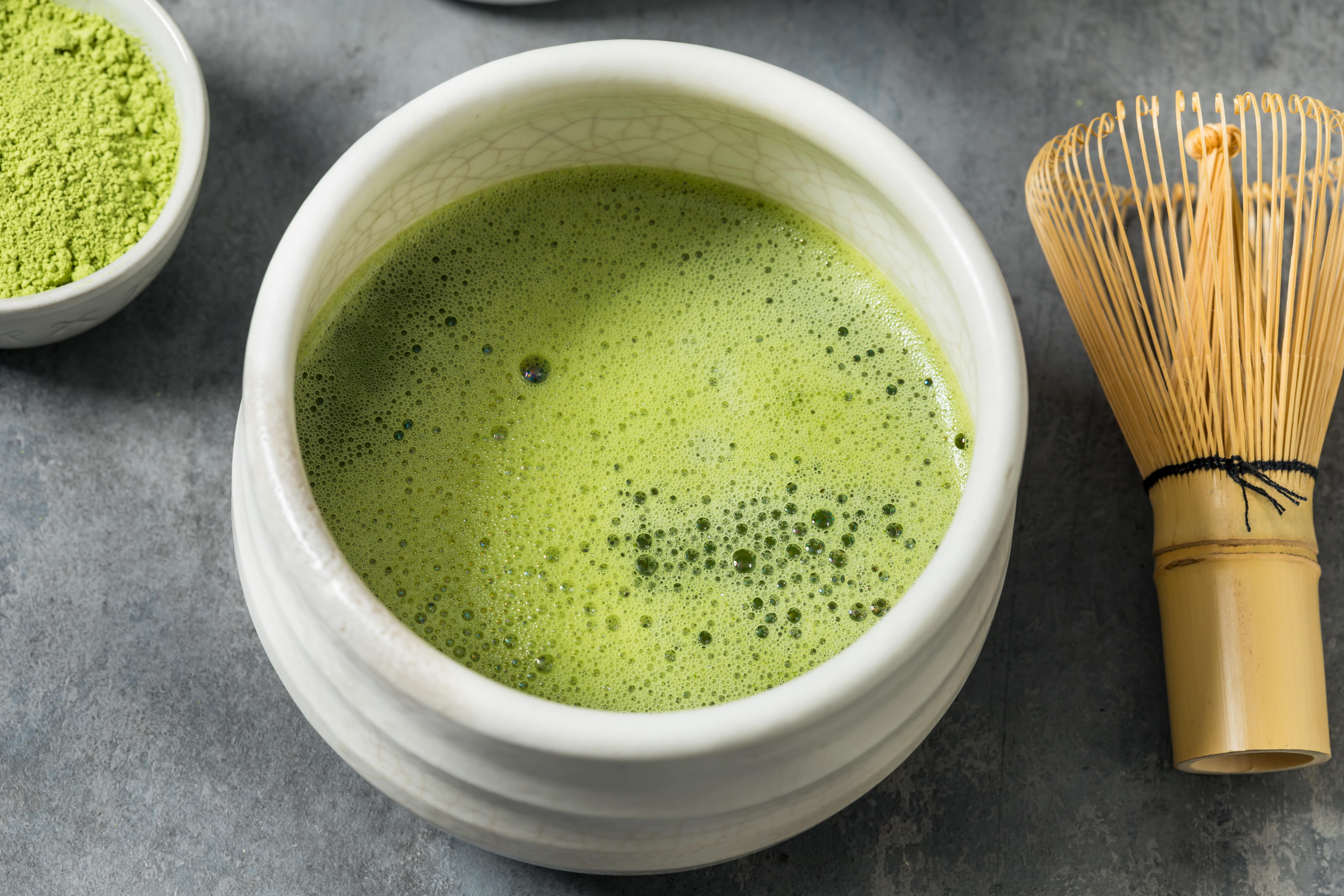 Matcha green tea in white bowl with powder and bamboo whisk beside it