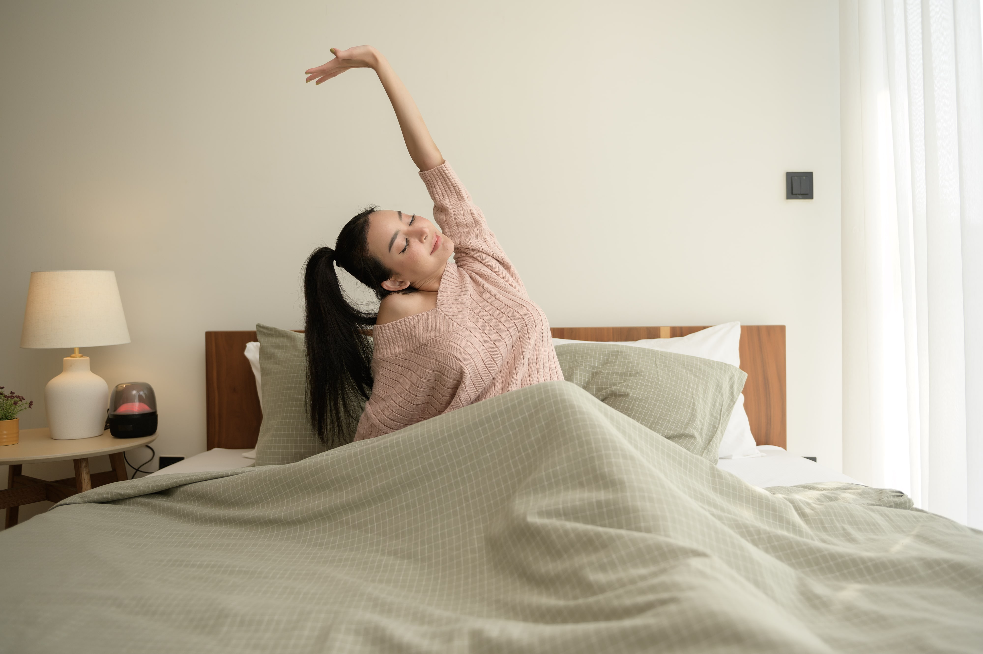 Woman stretching in bed wearing pink sweater in minimalist bedroom