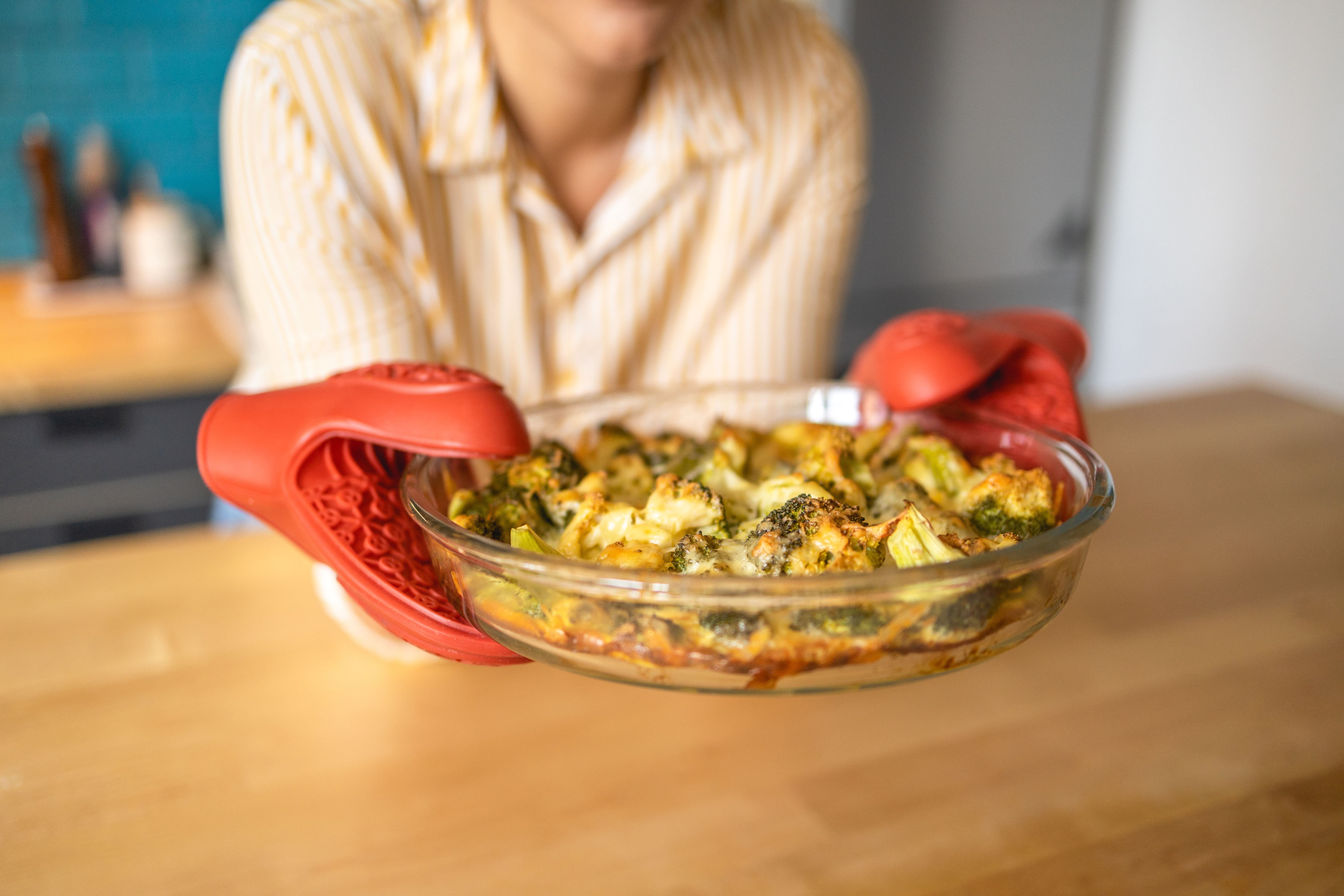 Woman in yellow striped shirt holding glass dish of roasted vegetables with red kitchen mitts