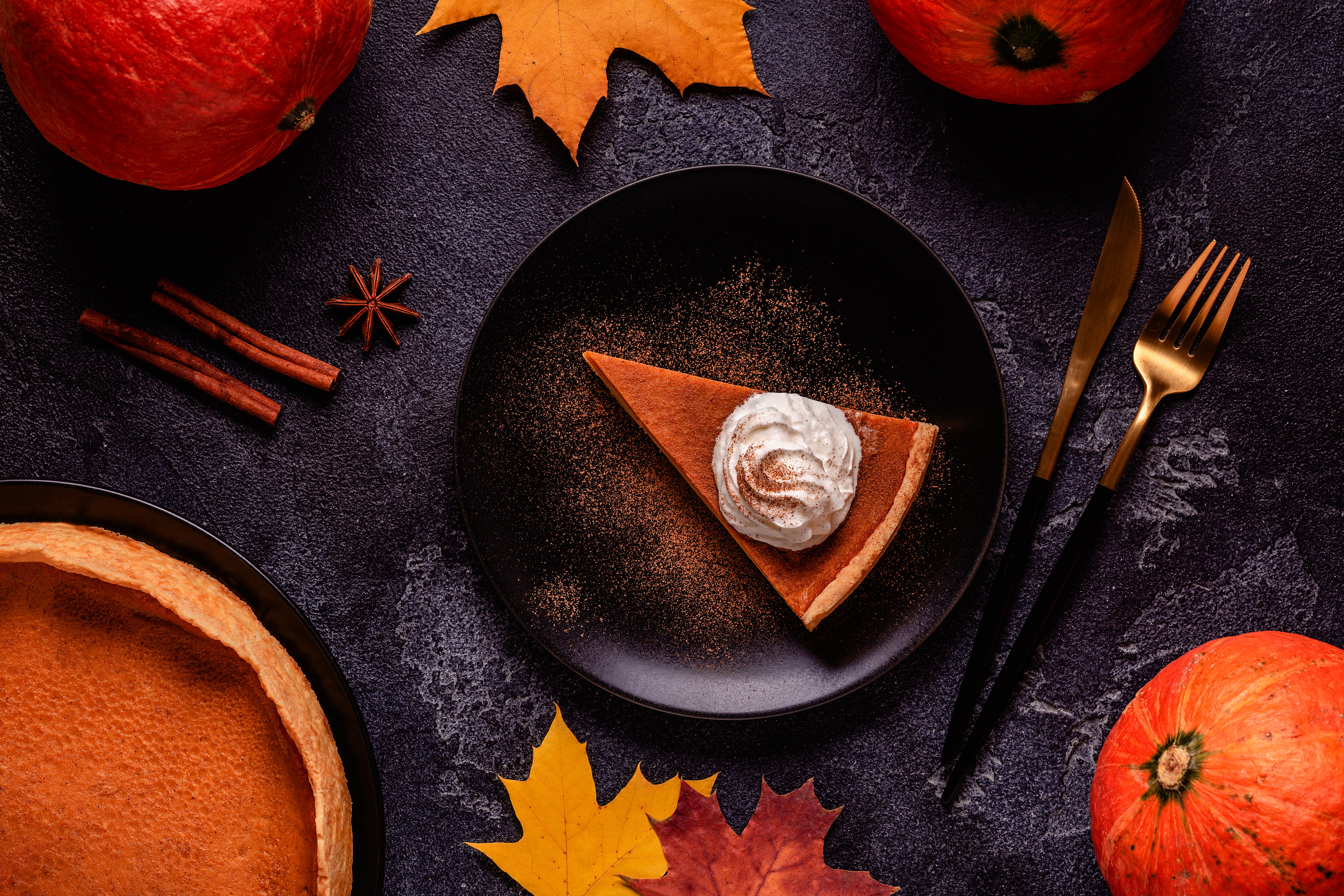 Autumn flatlay with pumpkin pie slice, whipped cream, fall leaves, pumpkins, and spices.