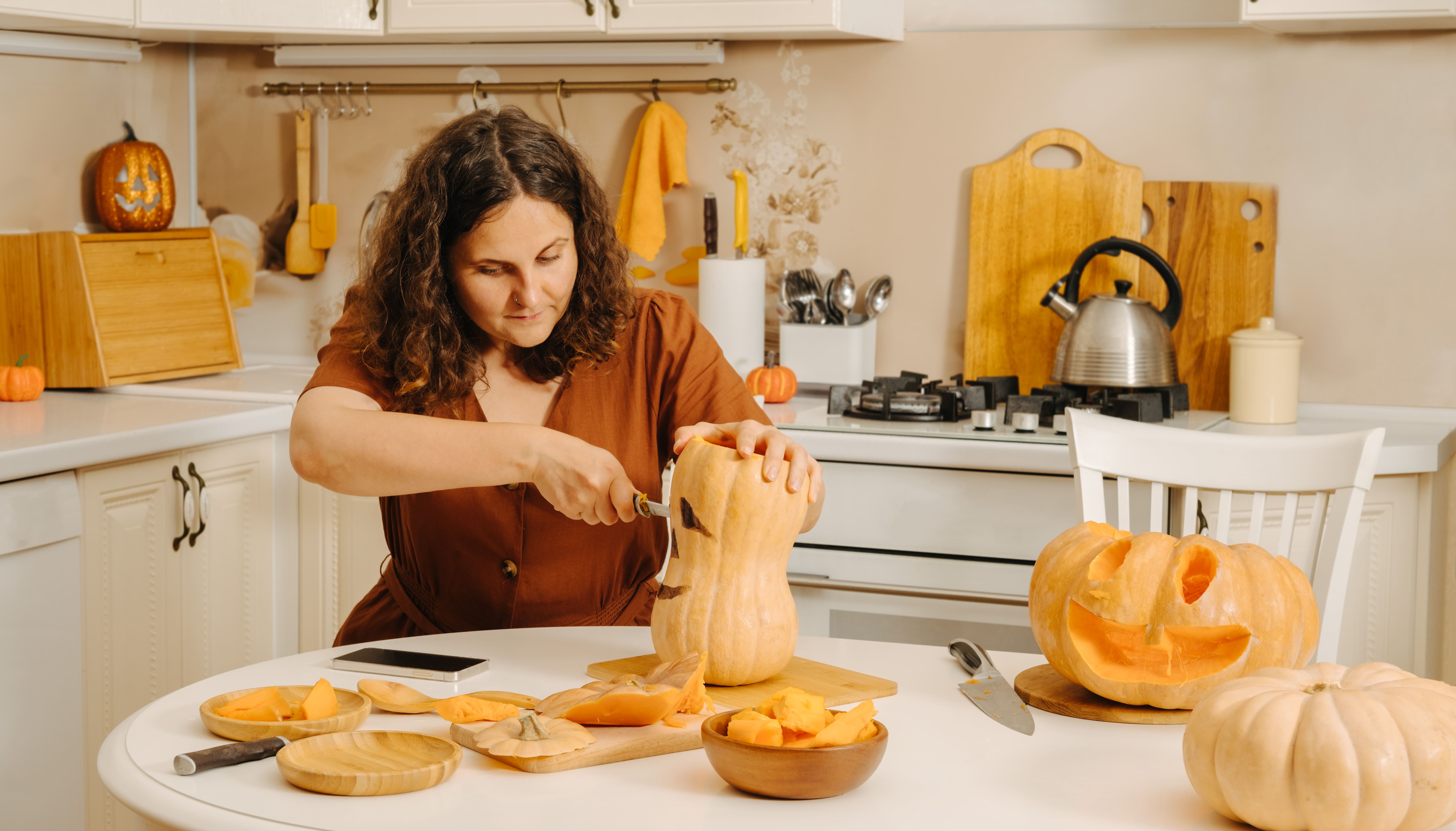 Woman carving pumpkins in a decorated kitchen during Halloween preparation.