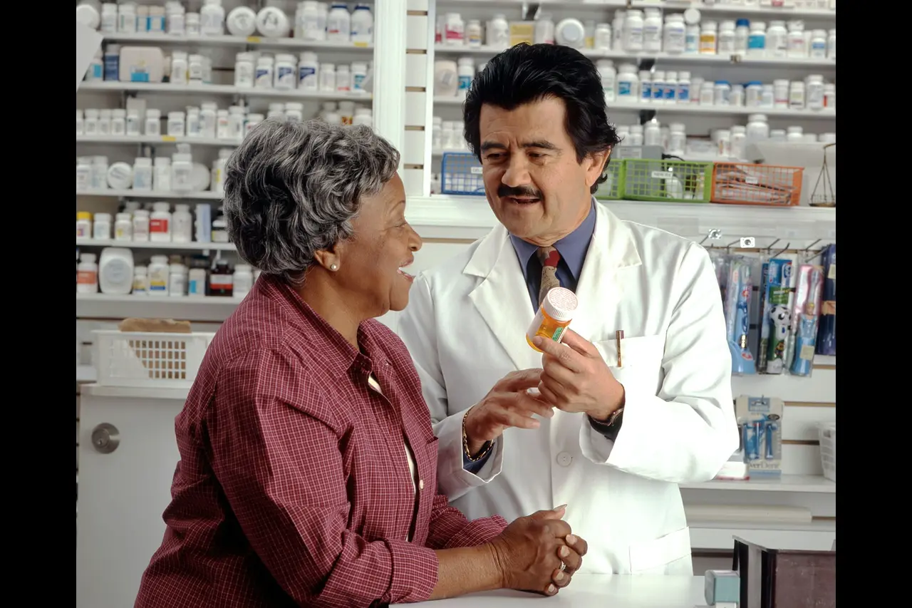 Pharmacist in white coat showing prescription medication bottle to senior woman customer in pharmacy