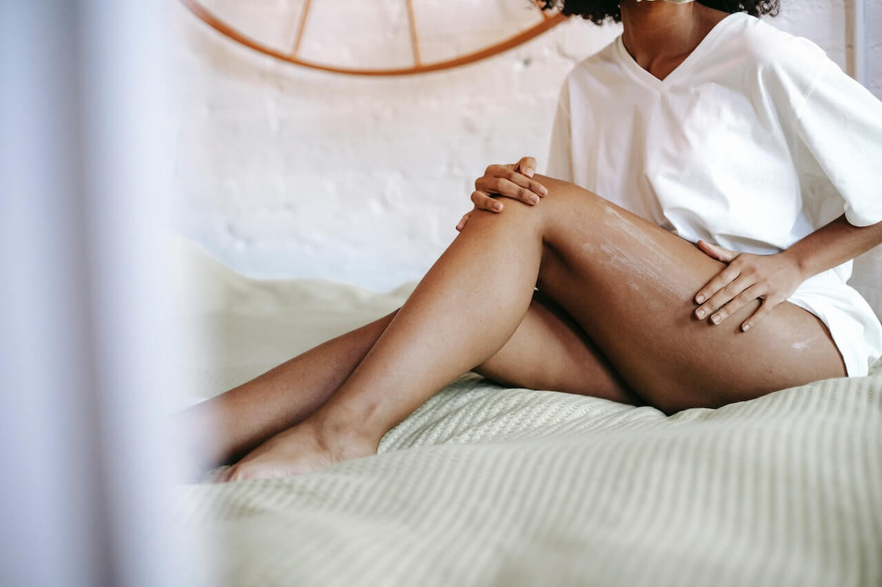 Woman applying skincare lotion to her legs in a bright bedroom