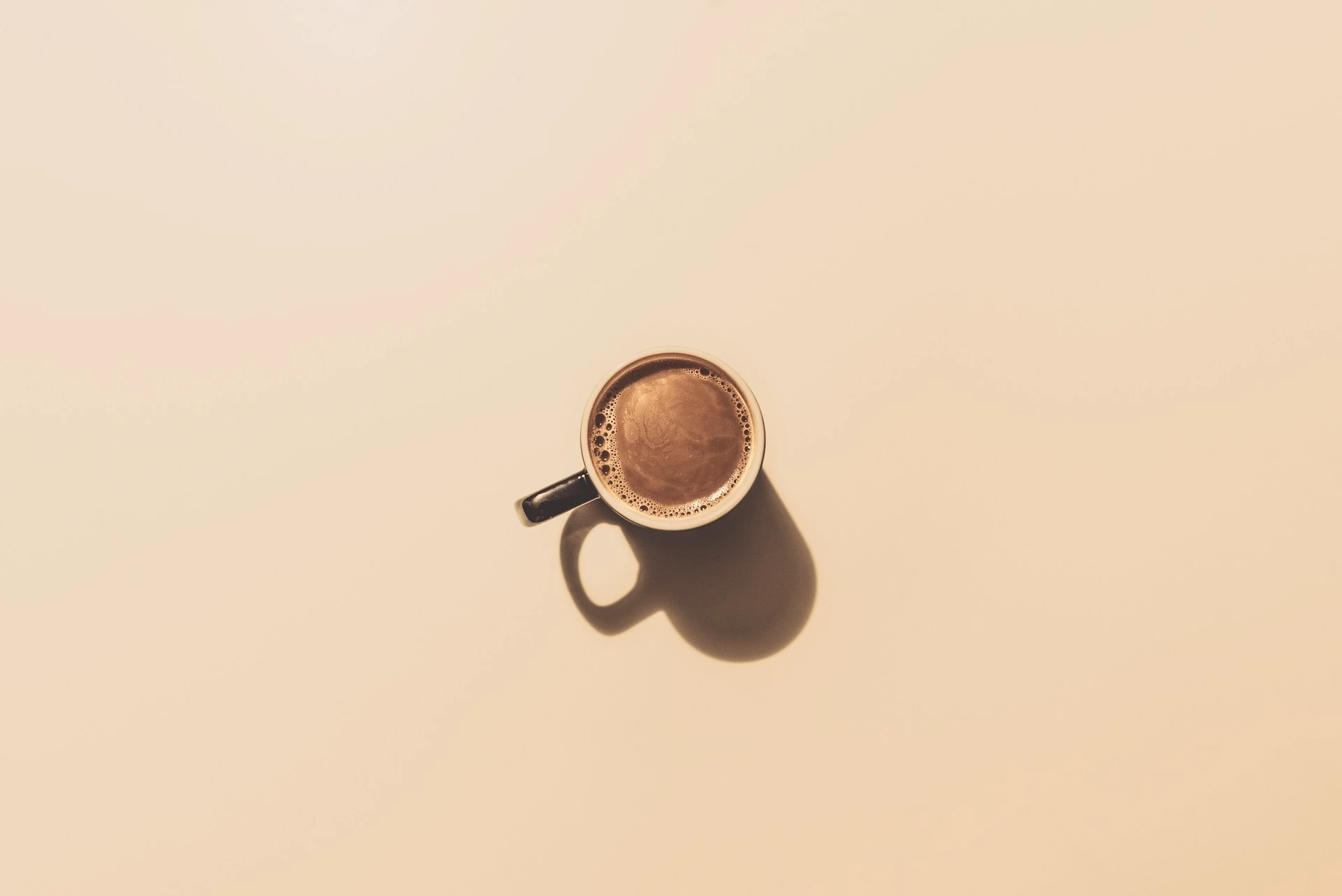 Top-down view of a dark mug with fresh coffee and foam on beige background.