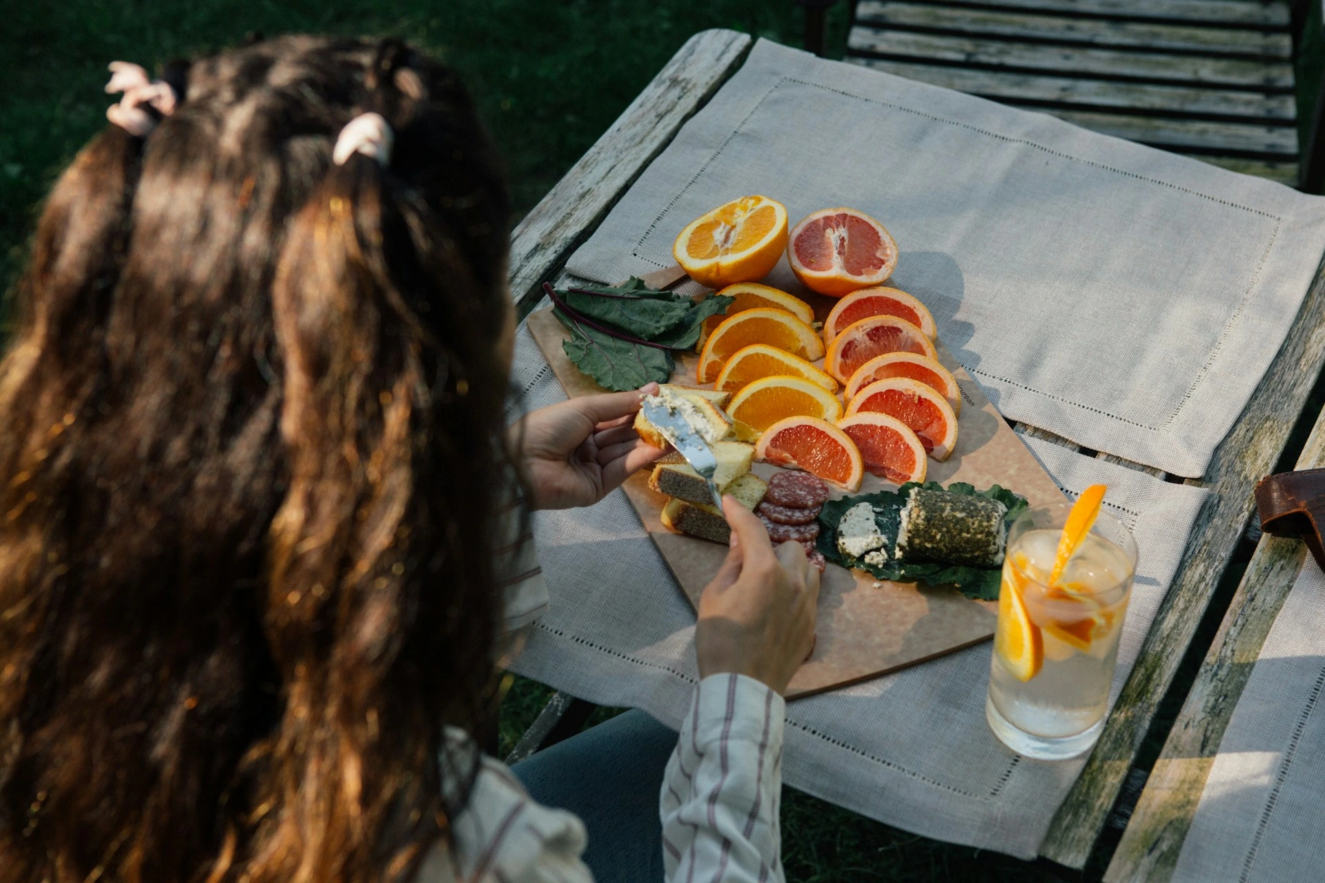Woman with long brown hair enjoys charcuterie and citrus fruits at outdoor wooden table